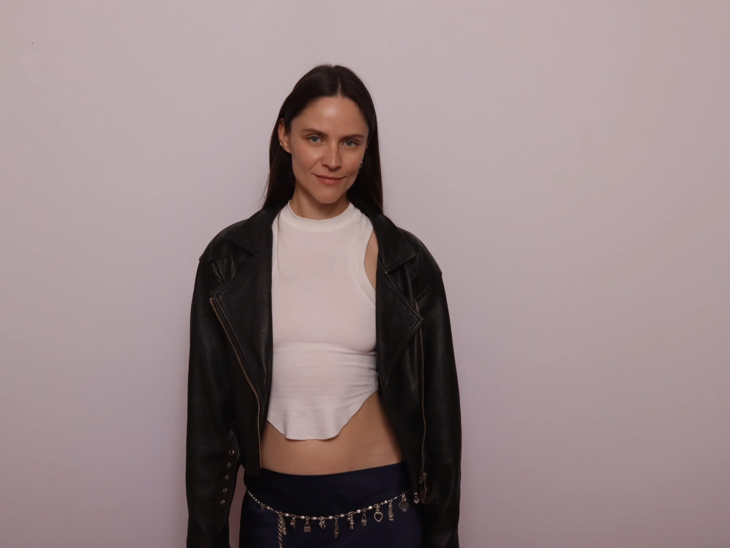 A woman with dark brown hair, light skin, and blue eyes standing against a plain white wall. She is wearing a black leather jacket over a white sleeveless top and navy pants with a silver chain belt.