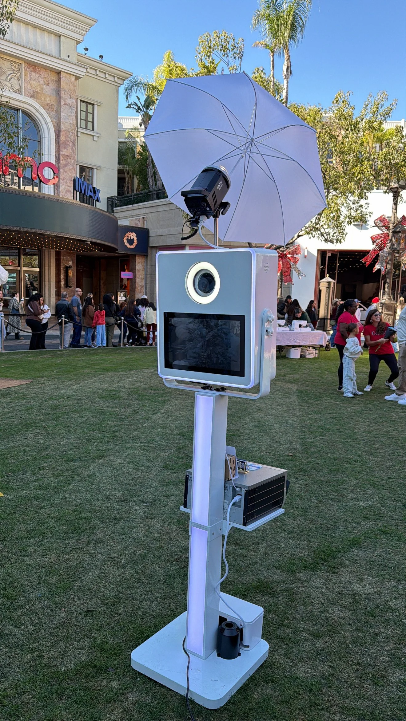 A photo booth set up outside at a public event, featuring a white frame with a camera lens, a screen, an umbrella for lighting, and a speaker, with people gathered in the background.