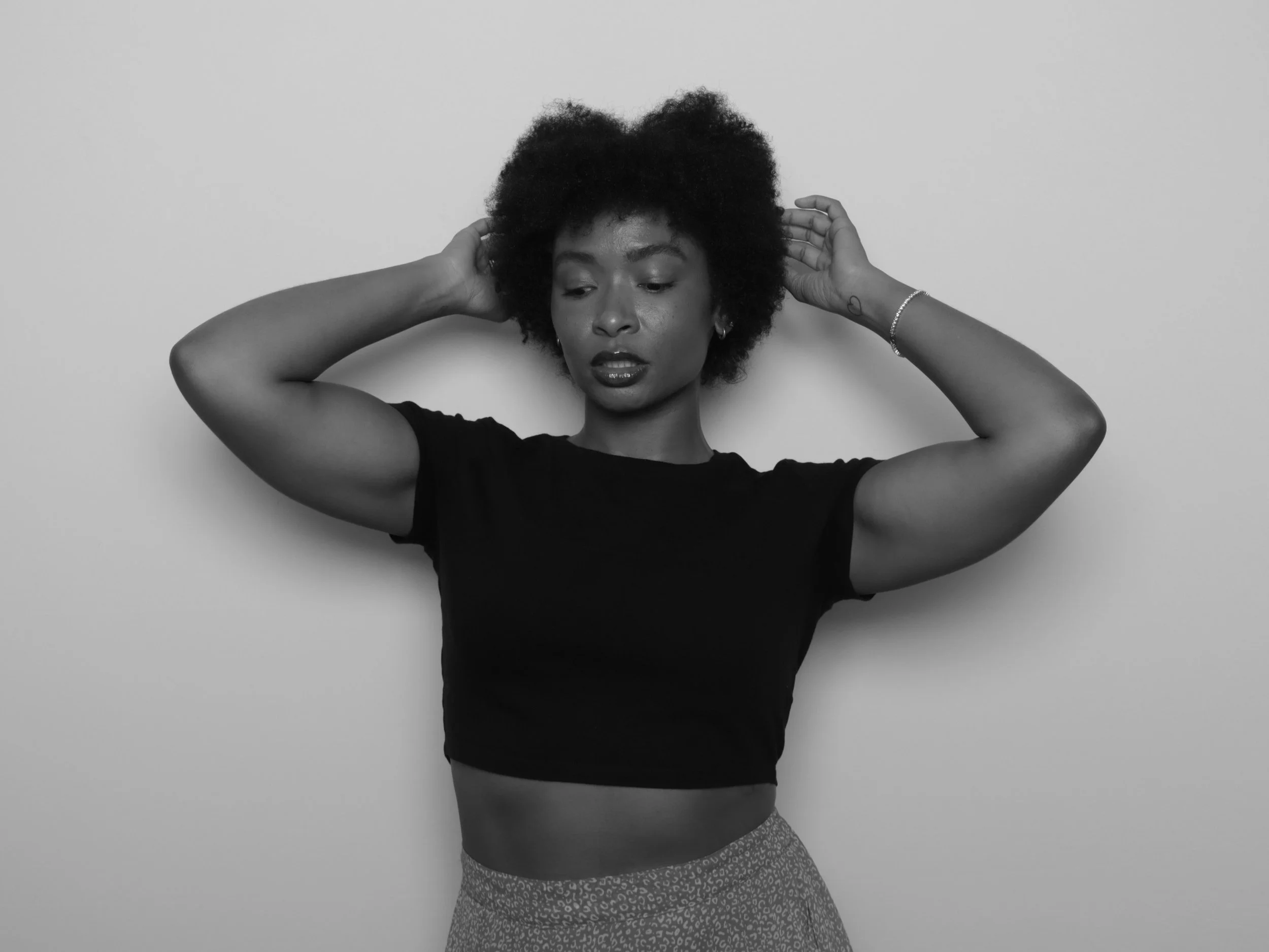 Black and white photo of a young woman with natural curly hair, wearing a black crop top and patterned pants, standing against a plain background, with her hands behind her head, looking down.