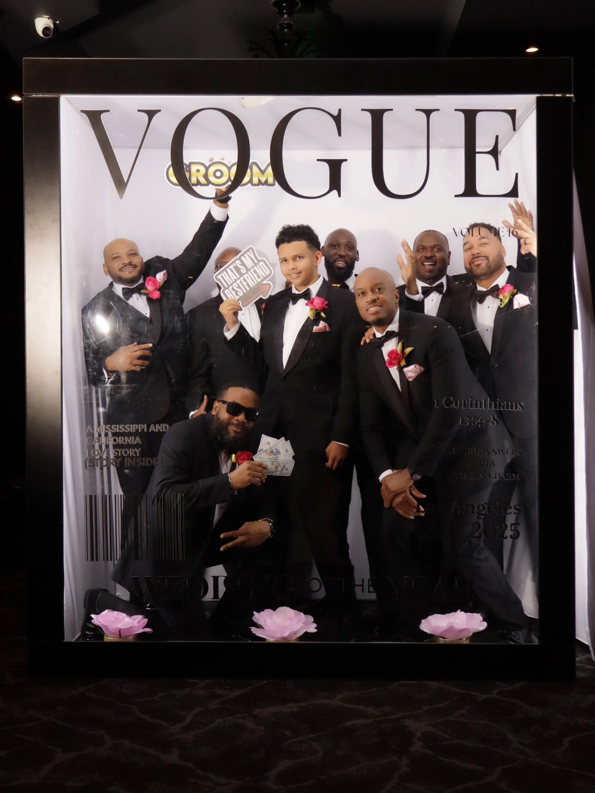 A group of men dressed in tuxedos celebrating inside a display frame resembling a Vogue magazine cover. They are smiling, some with sunglasses, and holding props like money and a sign that reads 'That's my best friend.' The display has large text rea