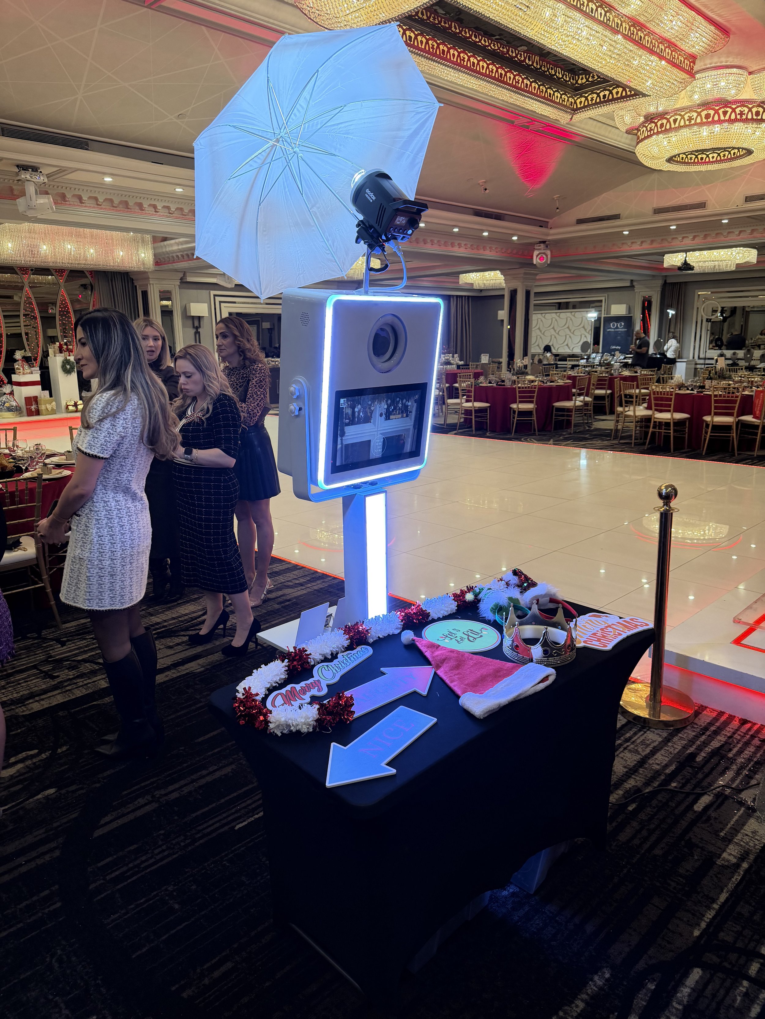 Photo booth with a camera, umbrella, and a display screen, set up for a Christmas celebration in an upscale banquet hall with people in the background.
