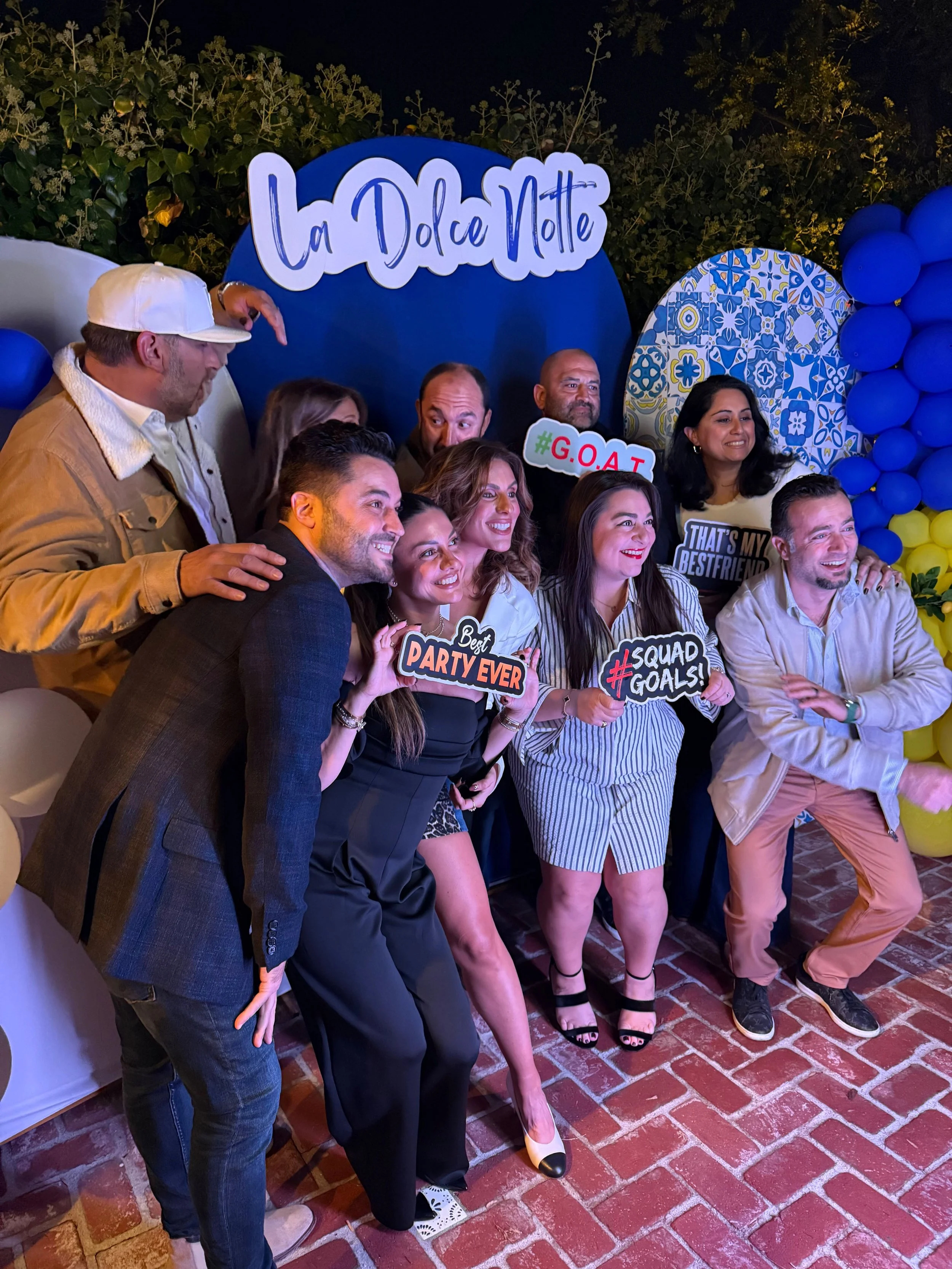 Group of nine people posing at a party, holding signs with phrases like "Best Party Ever,'' "#Squad Goals," and "#G.O.A.T," with a backdrop that says "La Dolce Notte" and decorative blue, yellow, and patterned balloons.