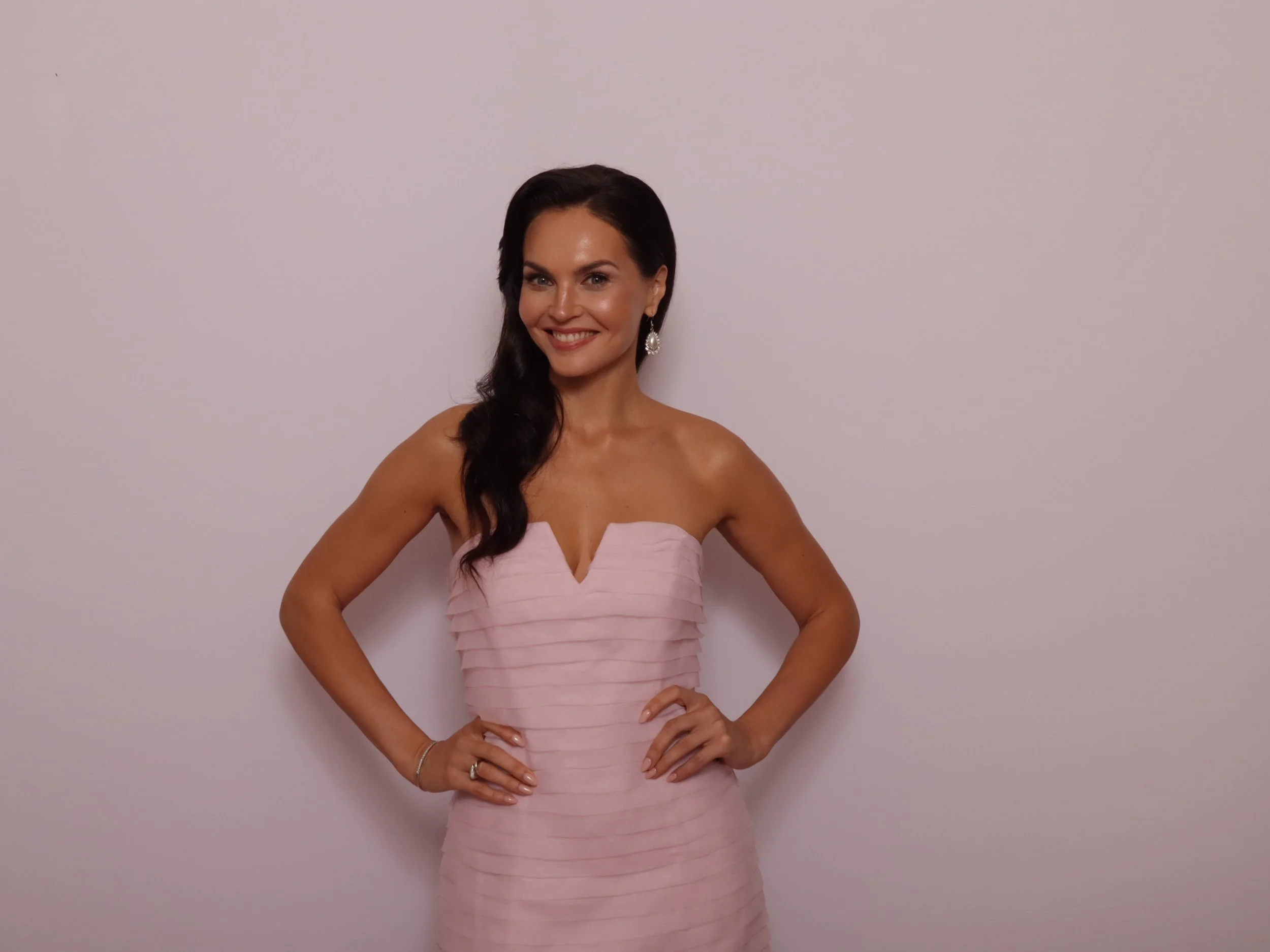 A woman with long dark hair, wearing a strapless pink dress with horizontal pleats, stands against a plain light-colored wall. She smiles and has her hands on her hips.