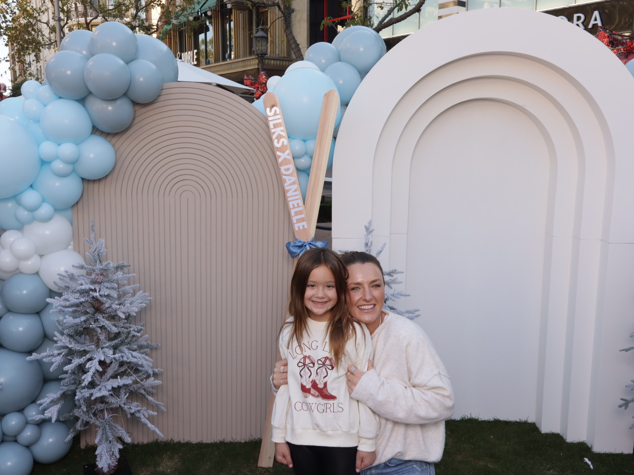 A woman and girl smiling together at a decorated outdoor event with white and blue balloon arrangements, small snow-covered trees, and a white arch backdrop.