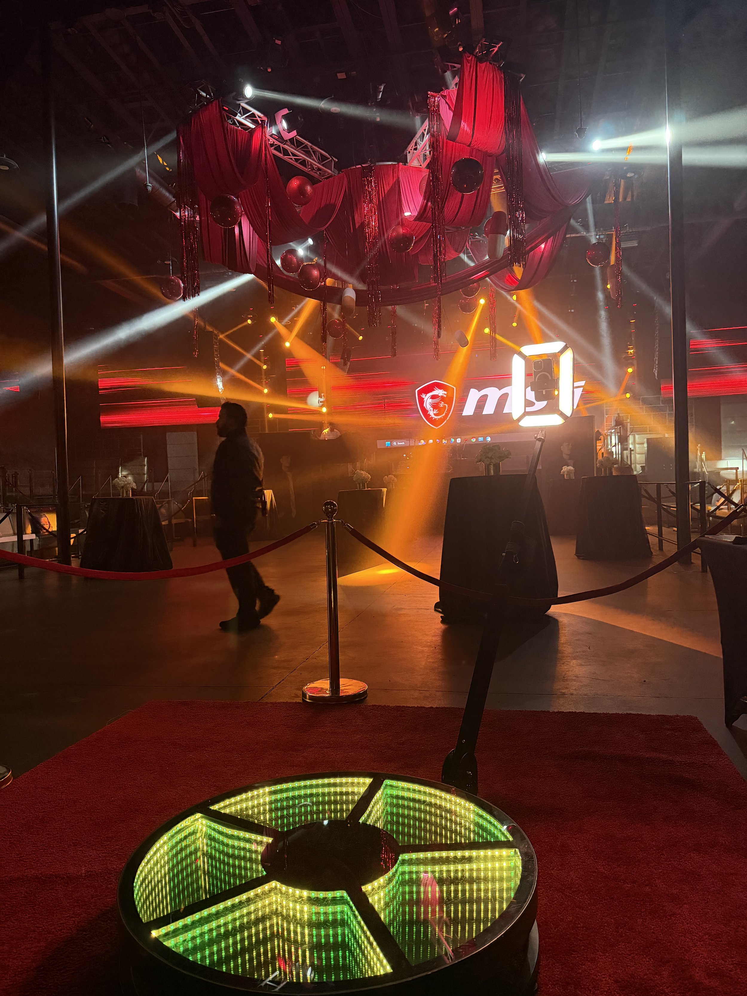 A nightclub or event venue with a stage decorated with red drapes and hanging red ornaments. There are colorful stage lights creating a lively atmosphere. The logo 'MSI' is displayed on a screen in the background. In the foreground, a lit circular pl