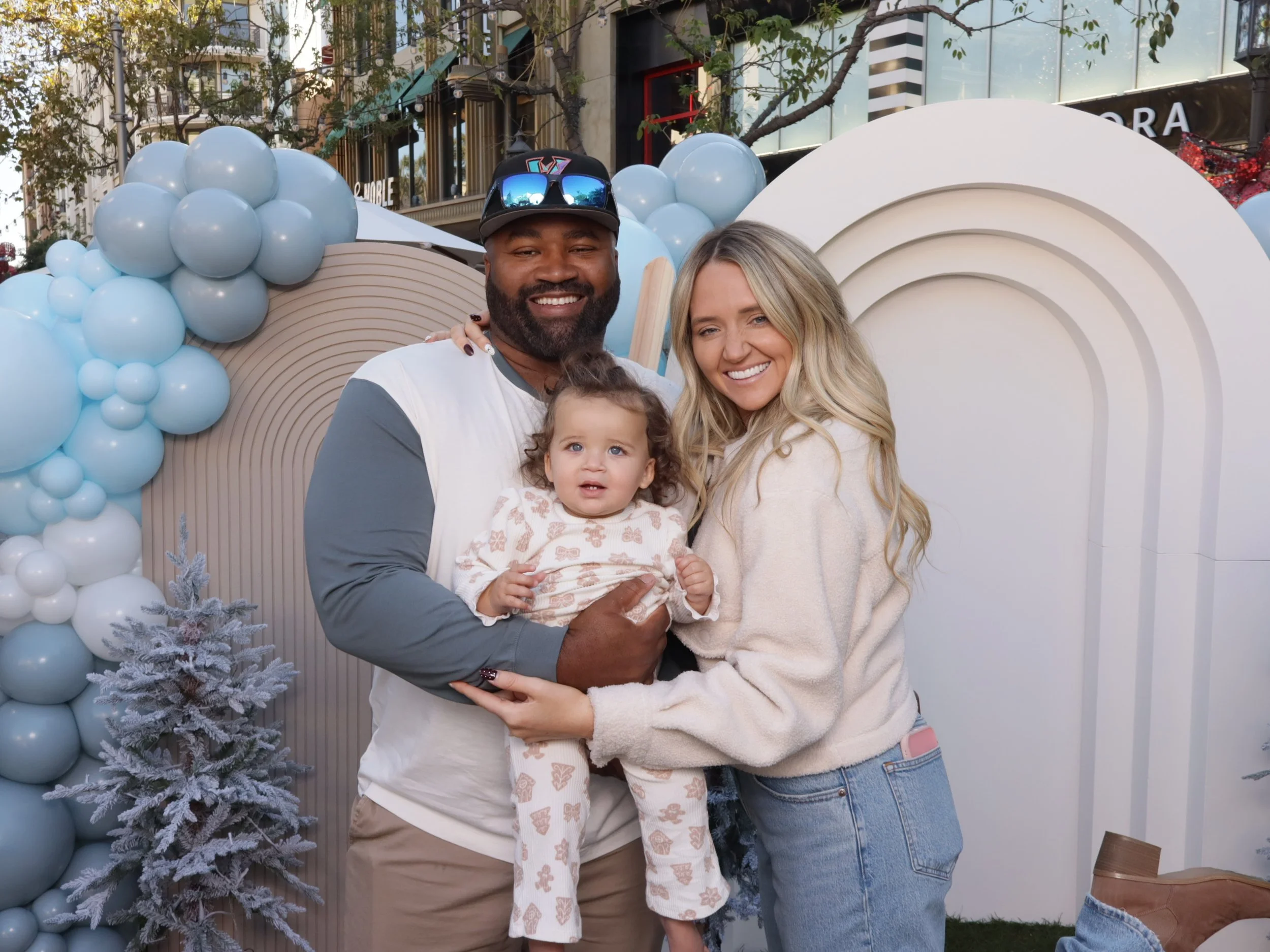 Smiling family of three, parents and young child, posing together outdoors at a festive event with a balloon and snowflake decoration backdrop.