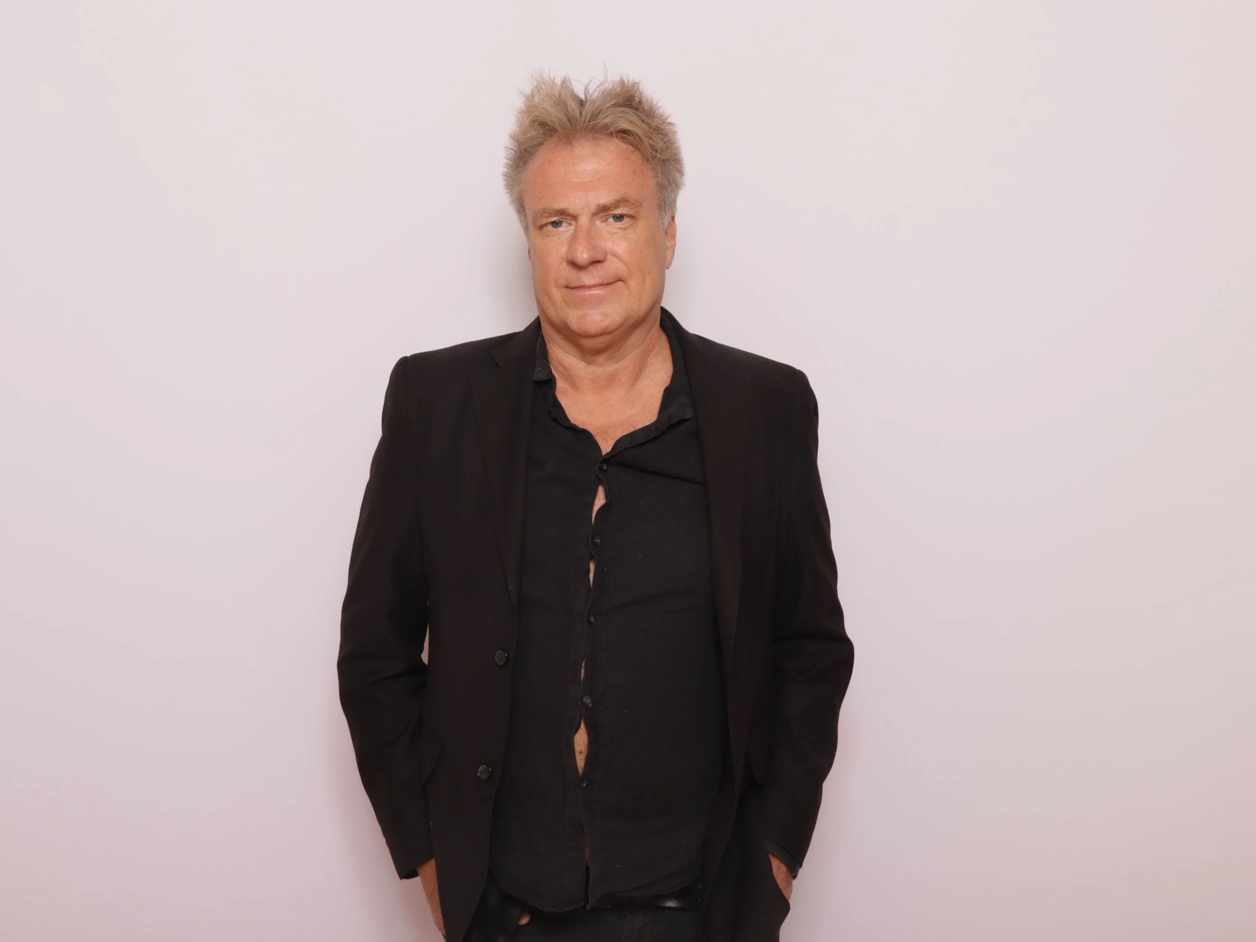 A middle-aged man with light-colored, messy hair, dressed in a black blazer and shirt, standing against a plain white wall, looking at the camera with his hands in his pockets.