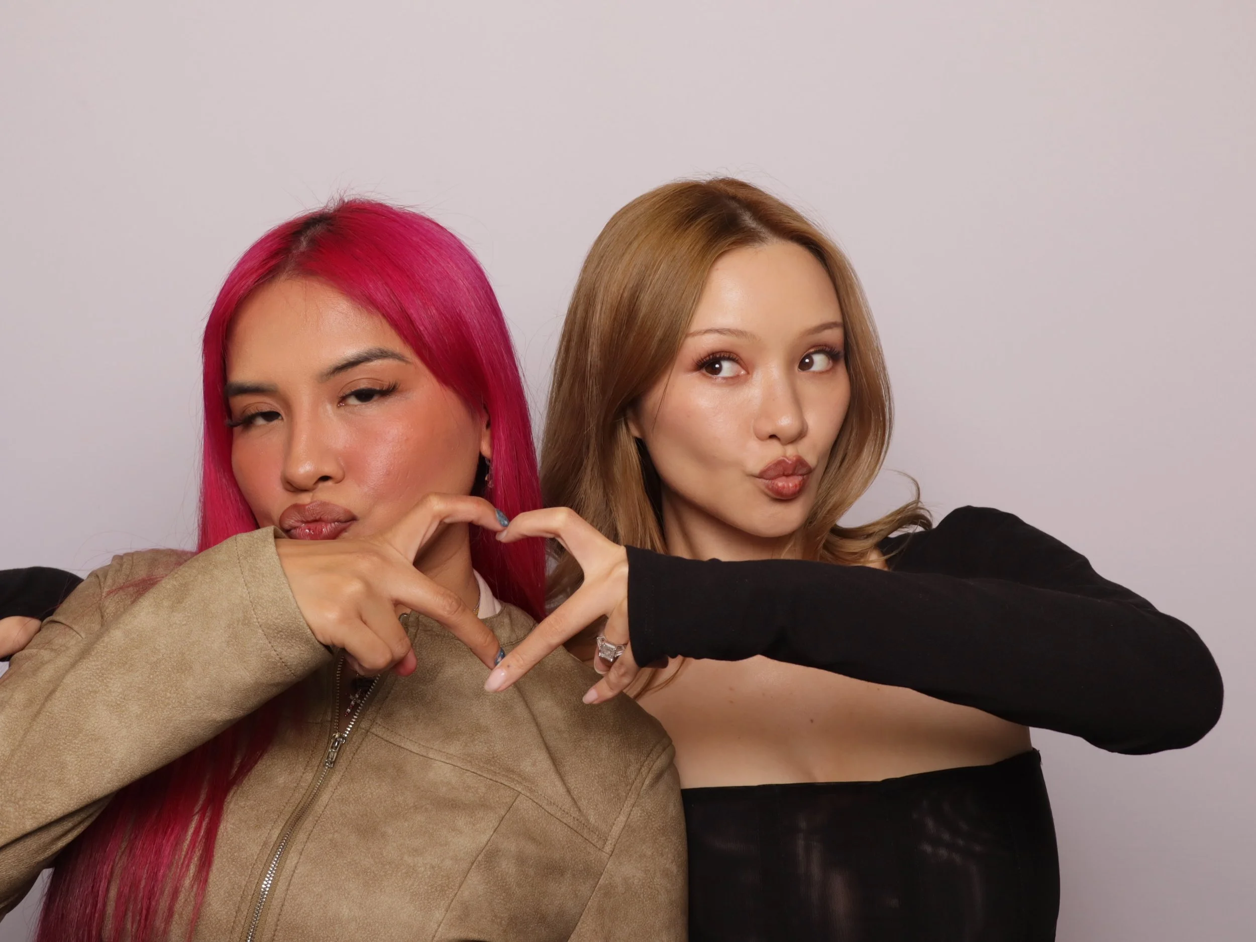 Two women posing together, making peace signs with their hands, one with pink hair and the other with blonde hair, against a plain background.
