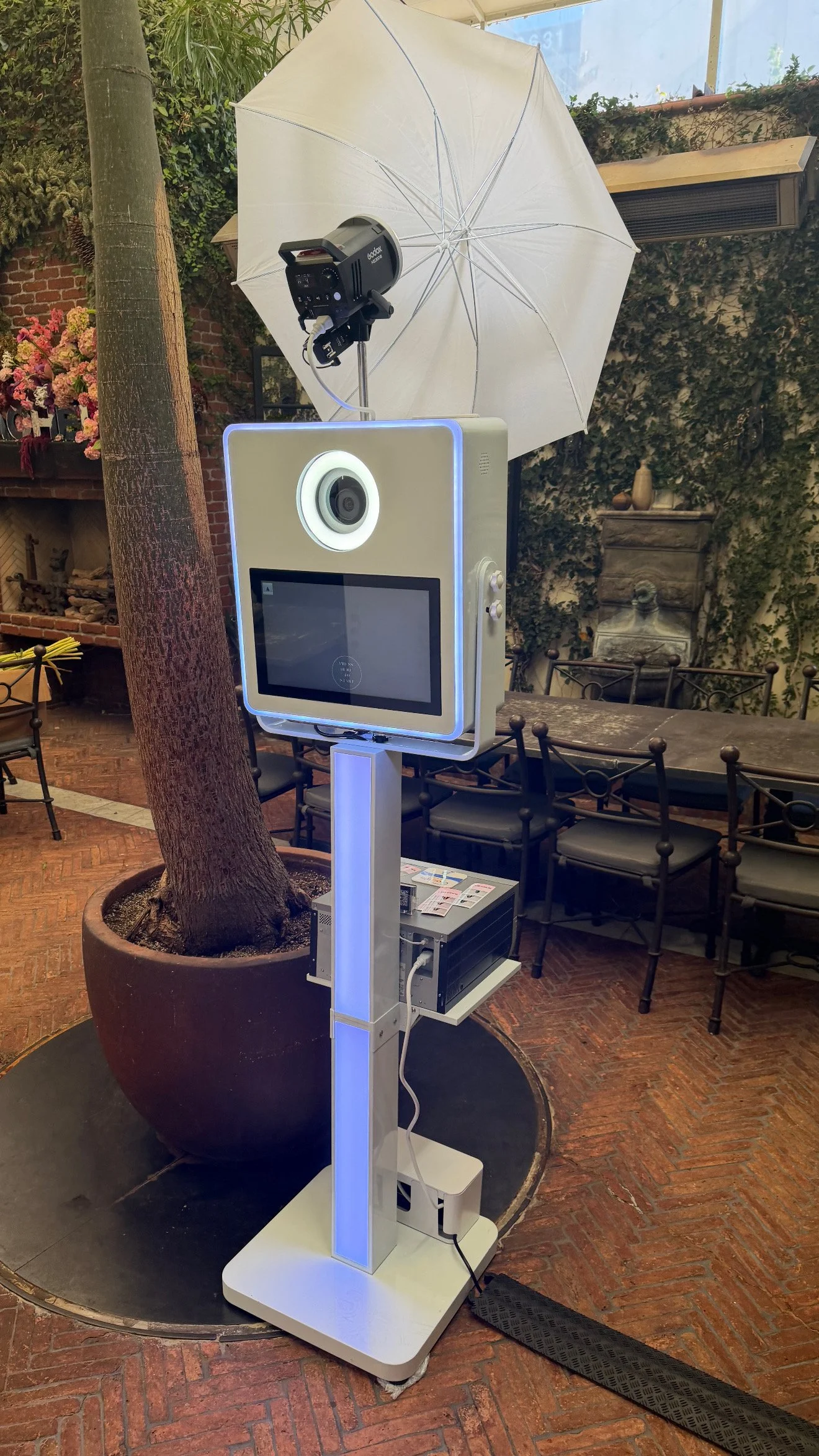 Photobooth setup with a large screen, camera, white umbrella for lighting, and various equipment inside a decorated indoor space with brick flooring and seating.