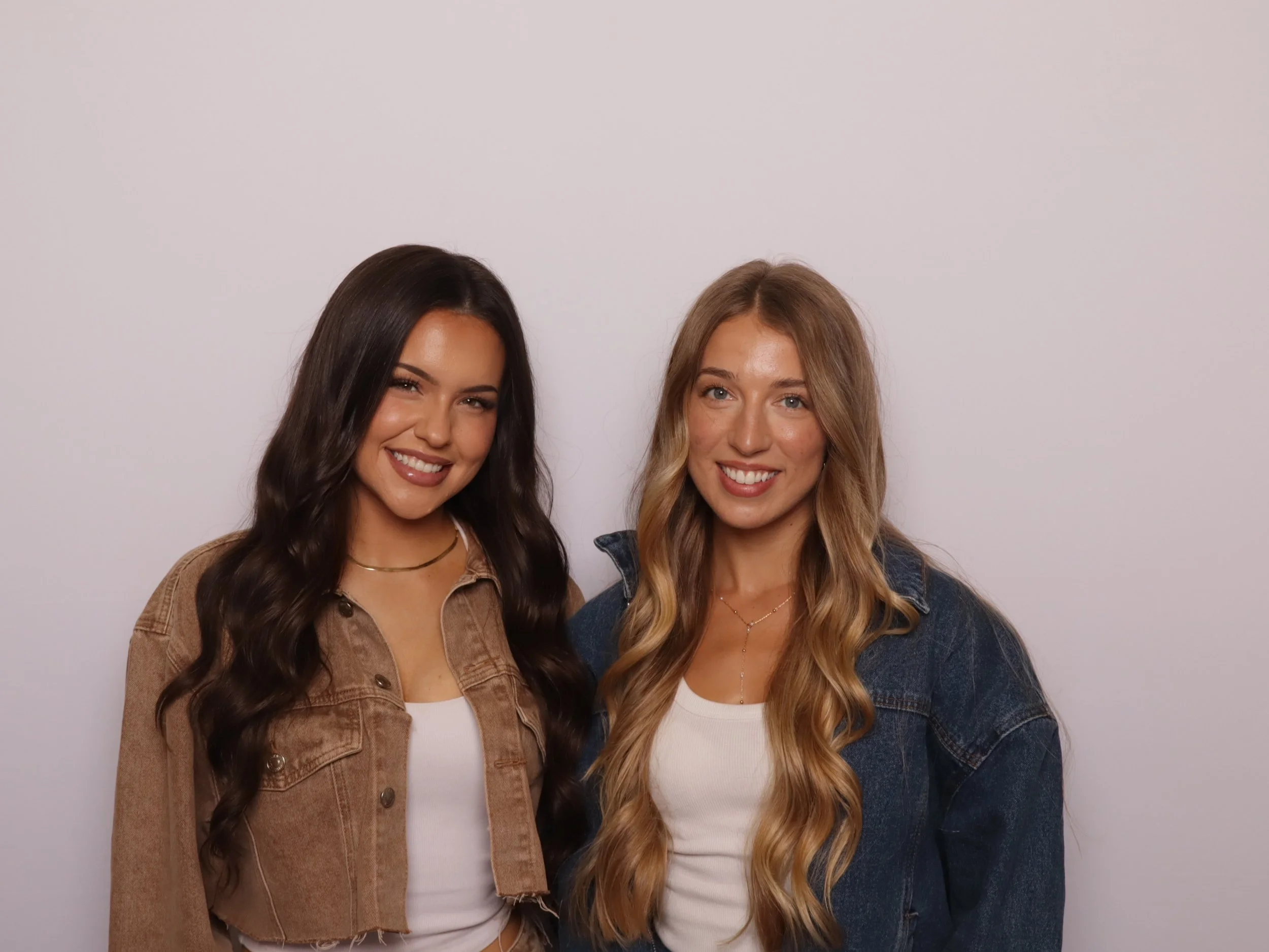 Two smiling women standing close together, one with dark wavy hair wearing a beige jacket and the other with blonde wavy hair wearing a denim jacket, in front of a plain light-colored background.