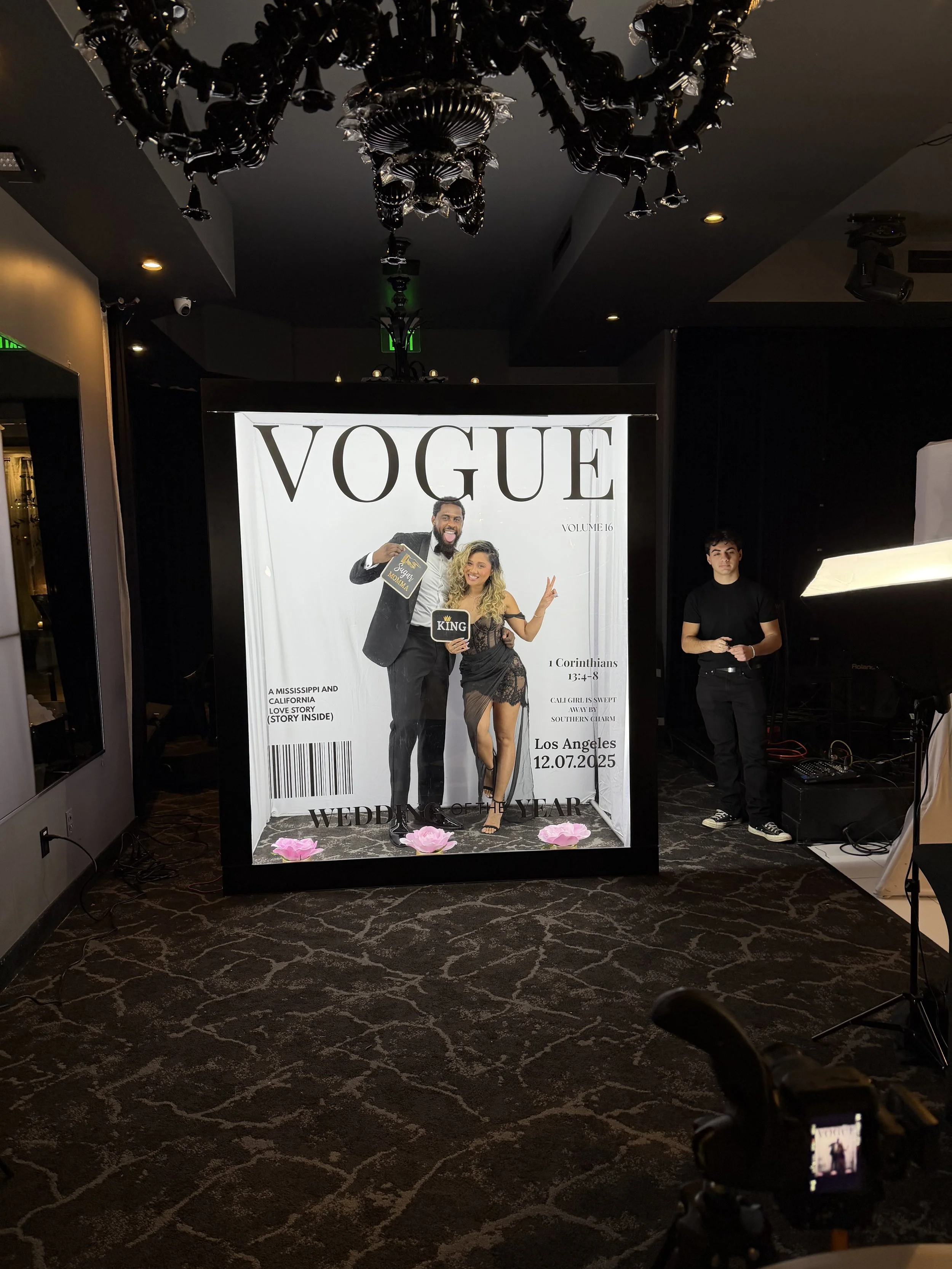 A photo of a photo booth at a Vogue event featuring a couple in formal attire, with the woman in a black dress and the man in a black tuxedo. The booth has printed text indicating it is for a wedding of the year event in Los Angeles on December 7, 20