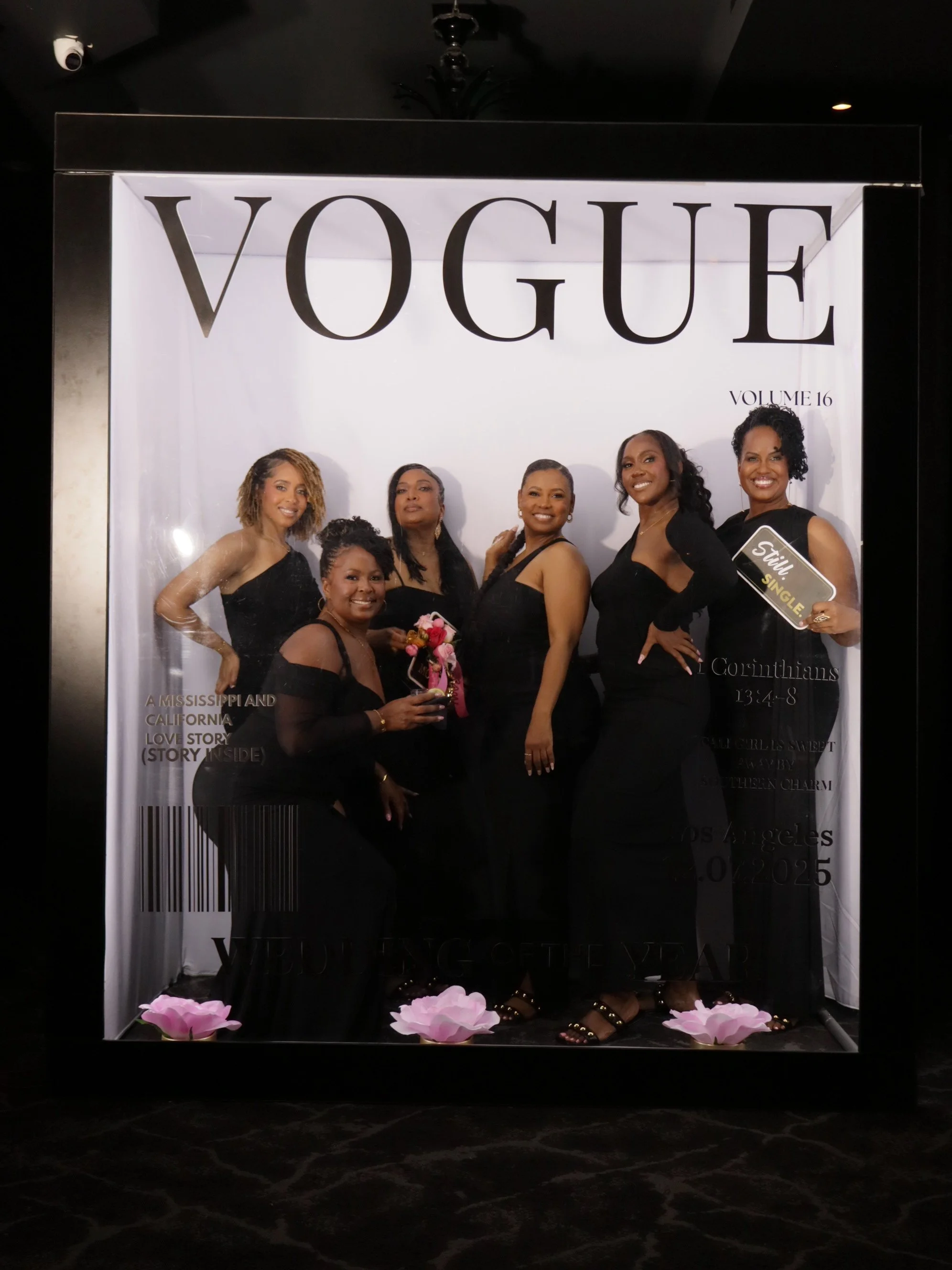 Six women dressed in black posing for a photo on a Vogue magazine-themed backdrop with large text, pink flowers on the floor, and some holding signs. The backdrop resembles a magazine cover.