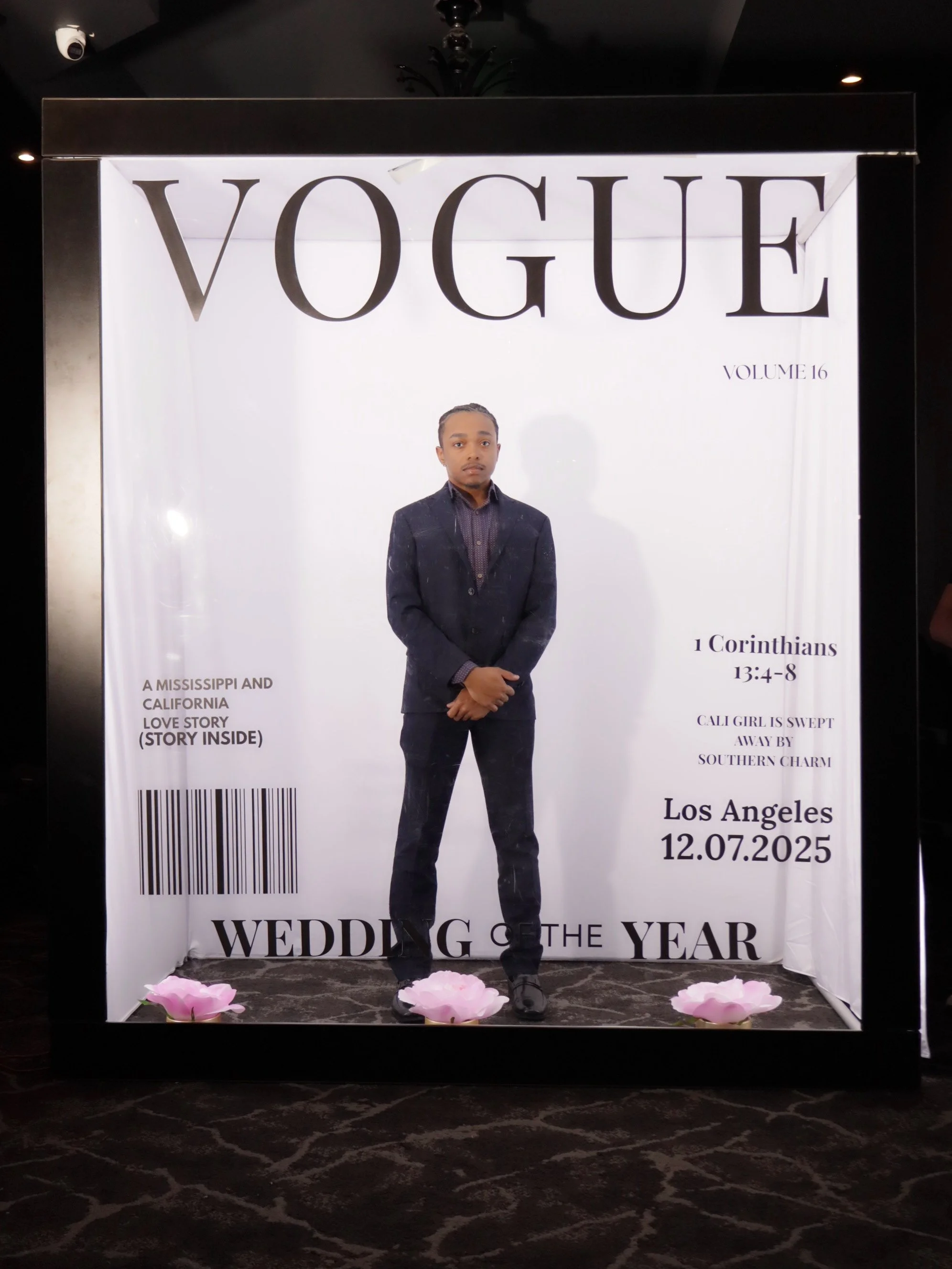 A person in a dark suit standing in front of a Vogue magazine cover display with the headline "Wedding of the Year," featuring the date 12.07.2025 and a Bible verse from 1 Corinthians 13:4-8.