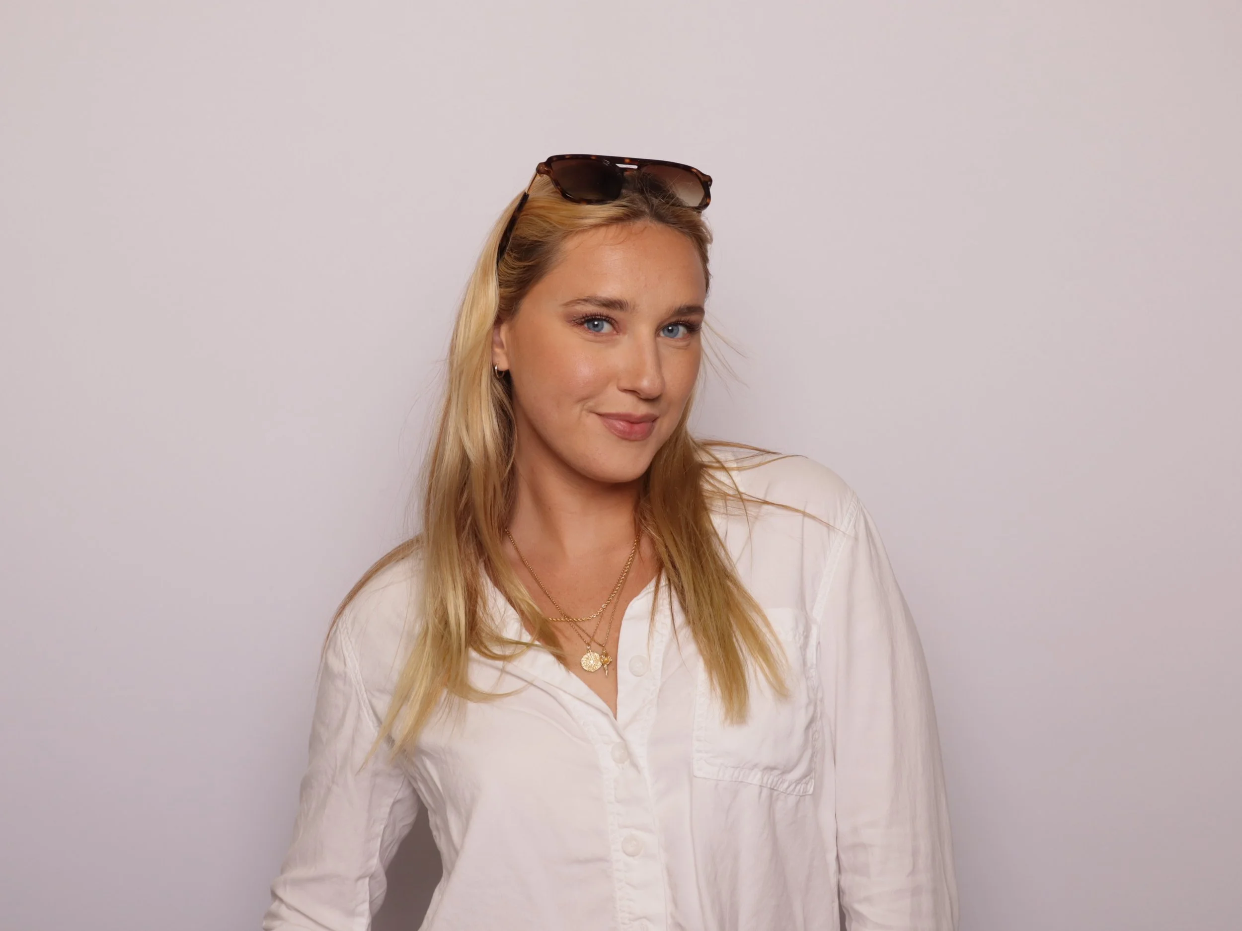 A young woman with long blonde hair, wearing sunglasses on her head, a white shirt, and layered gold necklaces, standing against a plain white background.