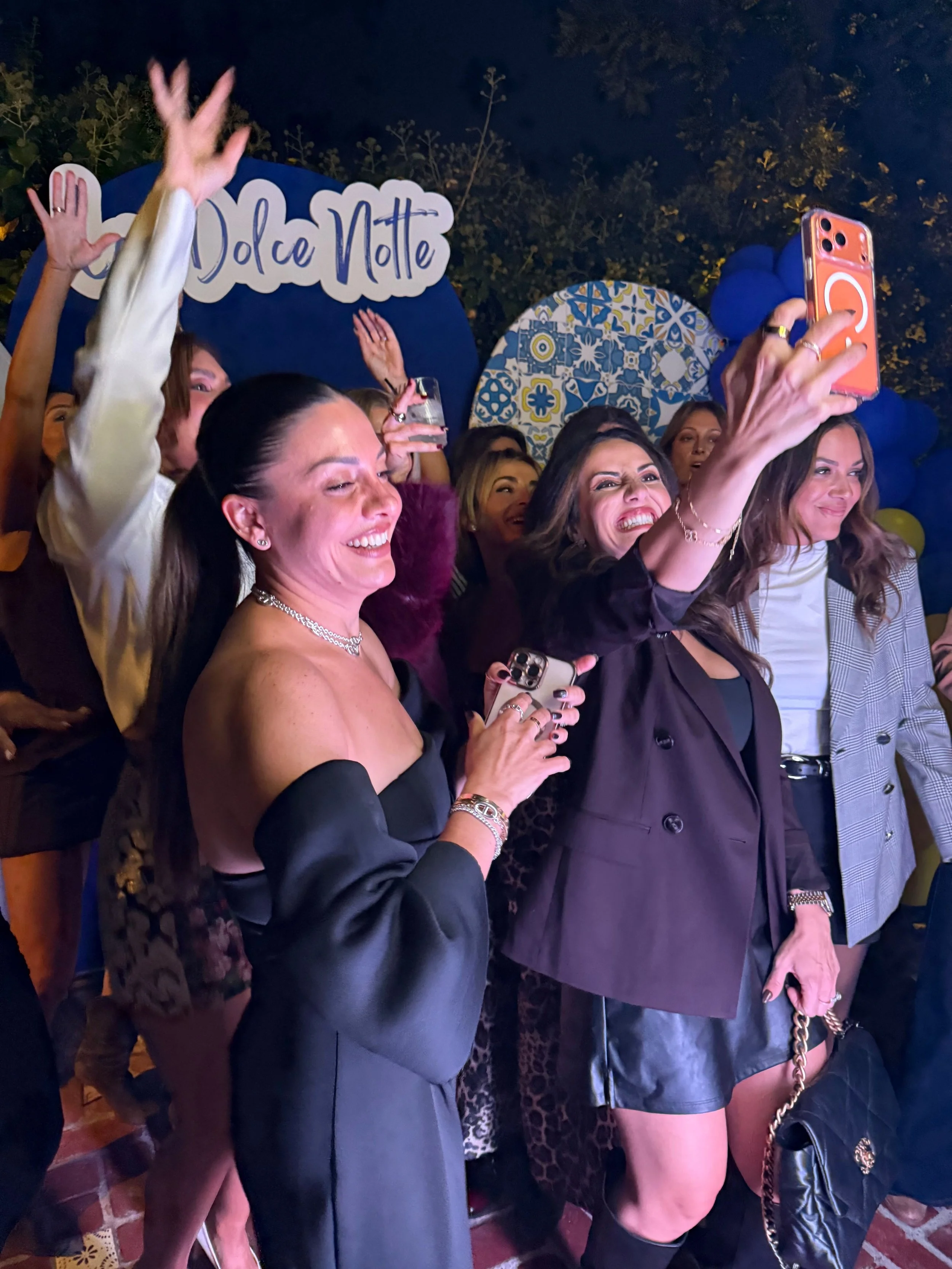 A group of women, some holding smartphones, taking selfies and smiling at a party with a Dolce Notte sign in the background.