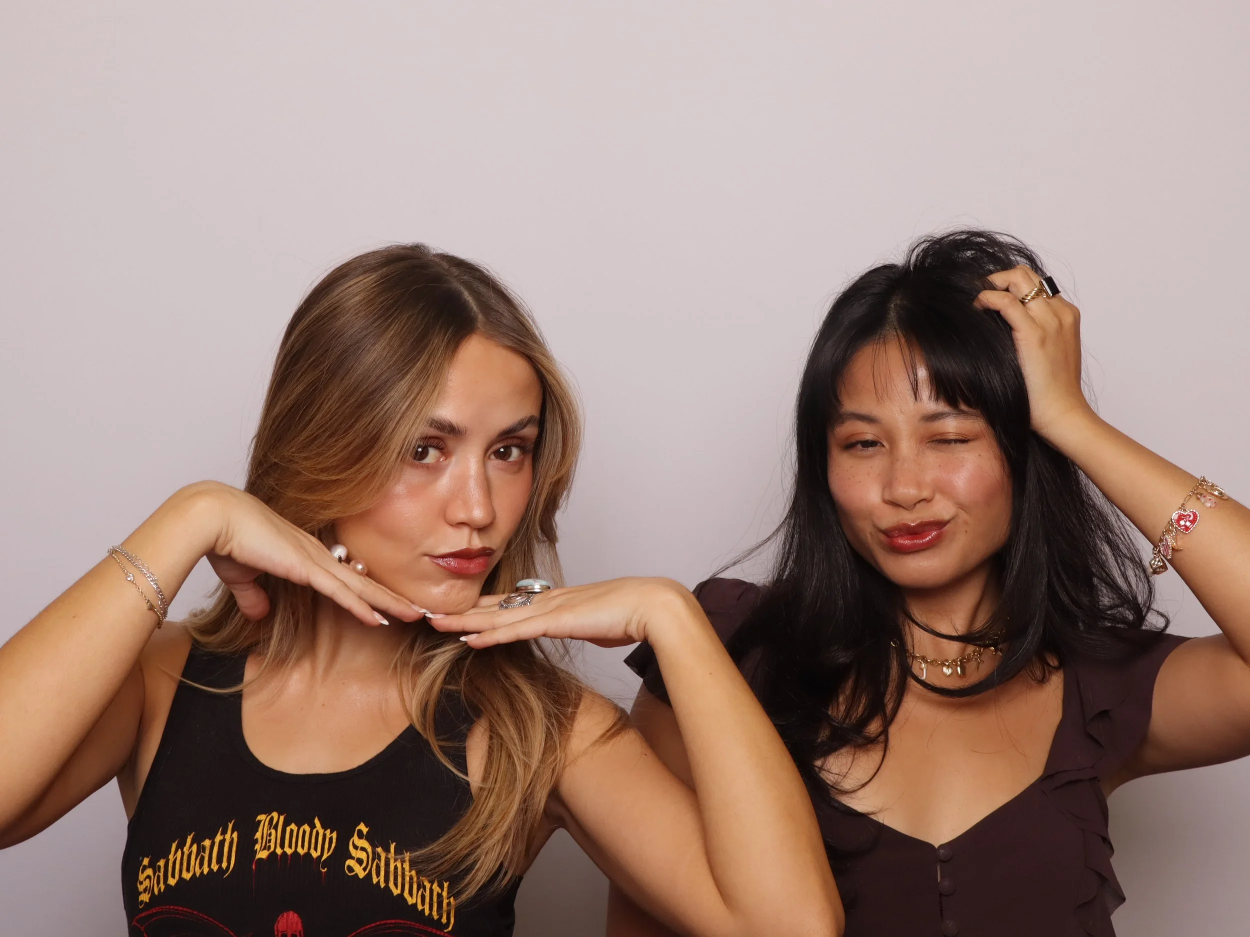 Two women posing playfully against a plain white background, one with blonde hair wearing a black Sabbath t-shirt, and the other with black hair wearing a dark top.