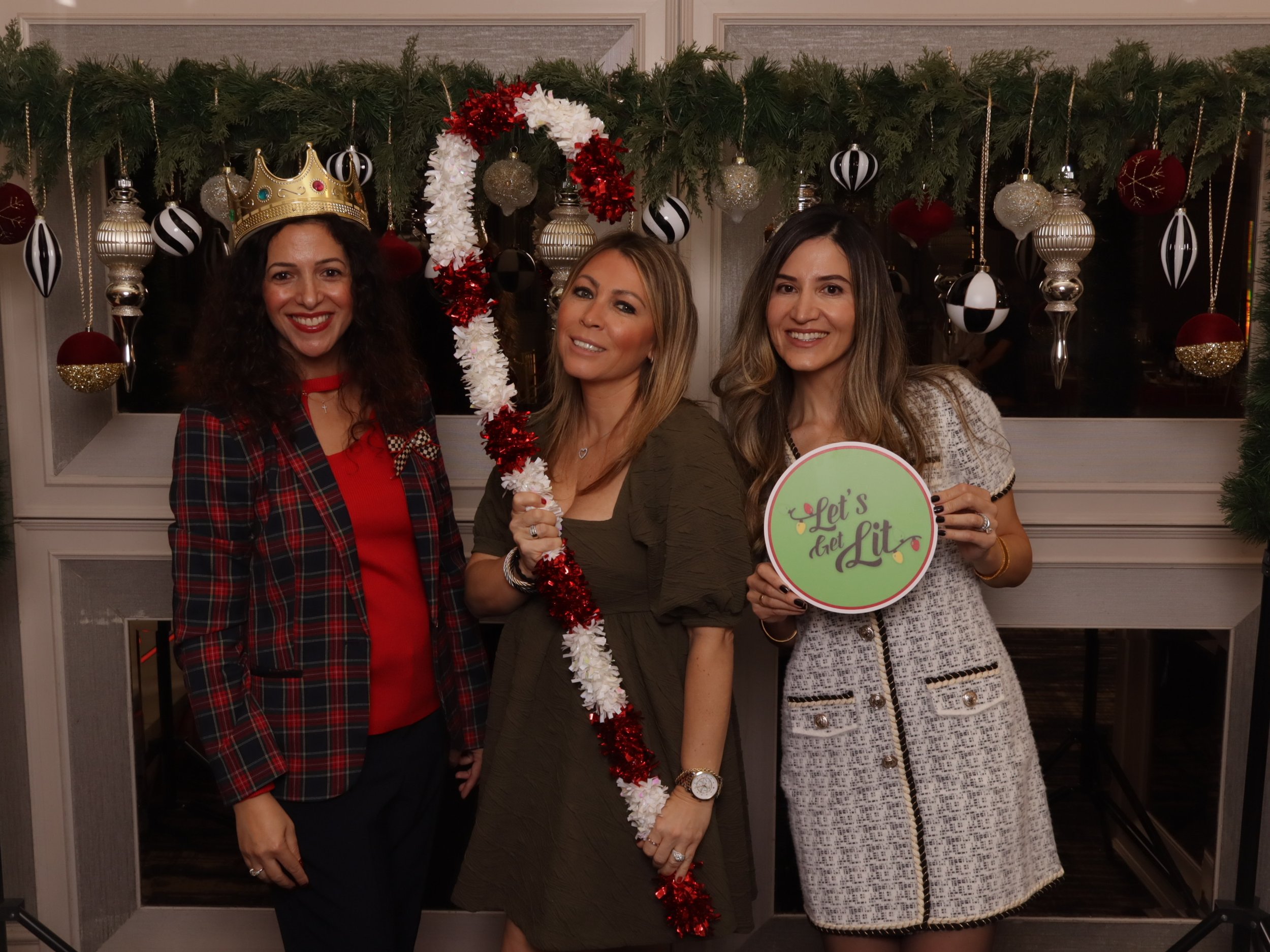 Three women standing in front of a decorated fireplace, smiling, during a Christmas celebration. The woman on the left wears a crown, red and plaid blazer, and red top; the woman in the middle holds a red and white garland and wears an olive-green dr