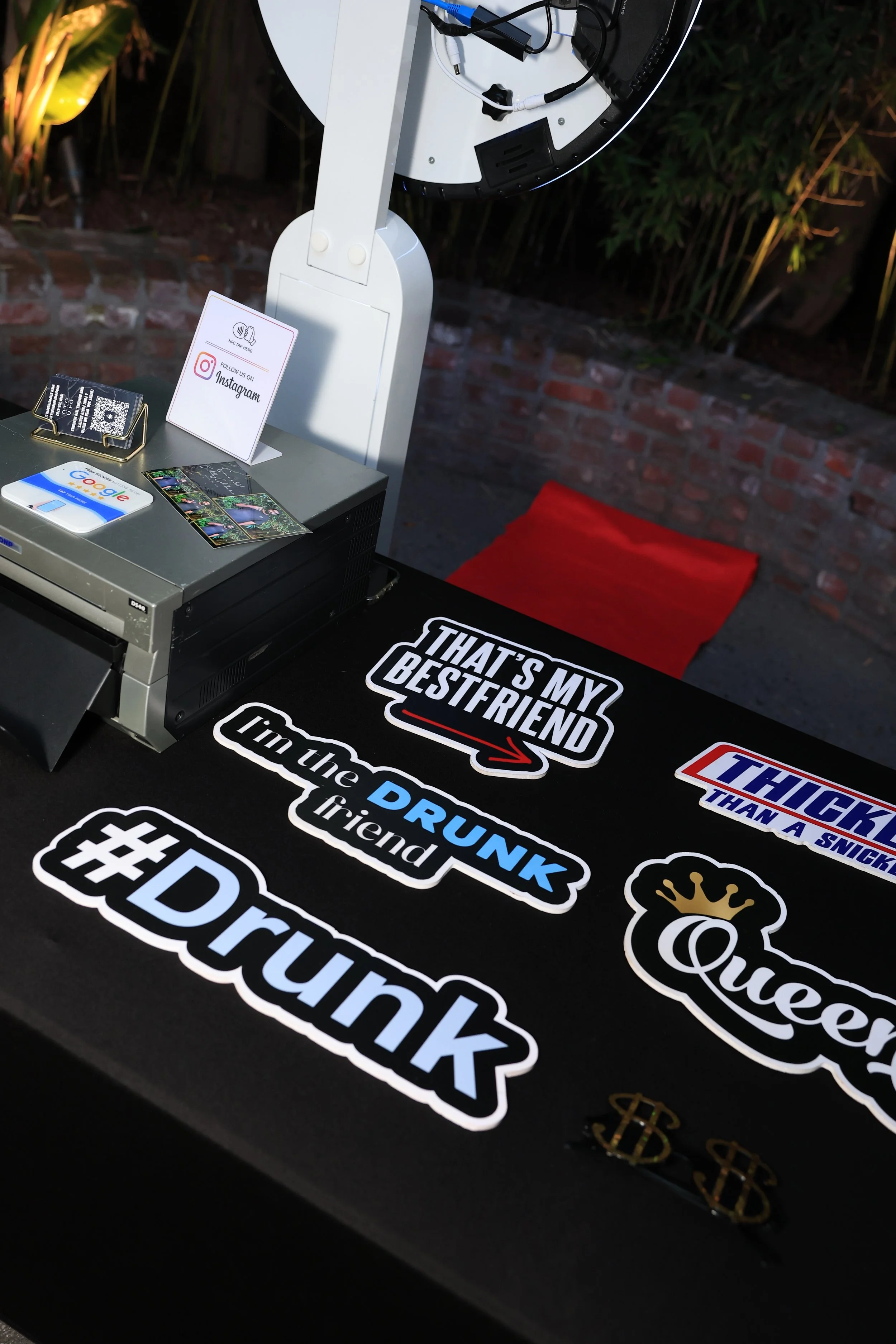 Table with humorous photo booth signs including "That's my best friend," "I'm the drunk friend," "#Drunk," "Queen," and "Check more than a snack." in front of a photo booth with props and a brick wall in the background.