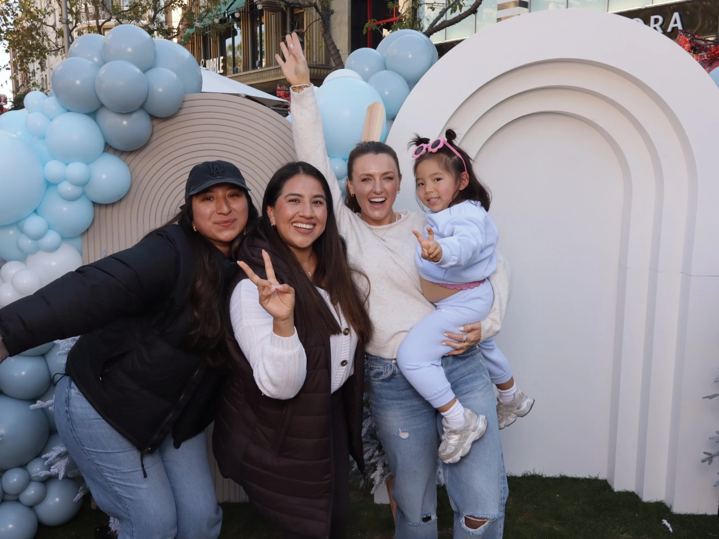Four women and a young girl pose together outdoors in front of a decorative background of blue balloons and a white rainbow-shaped structure, smiling and making peace signs.