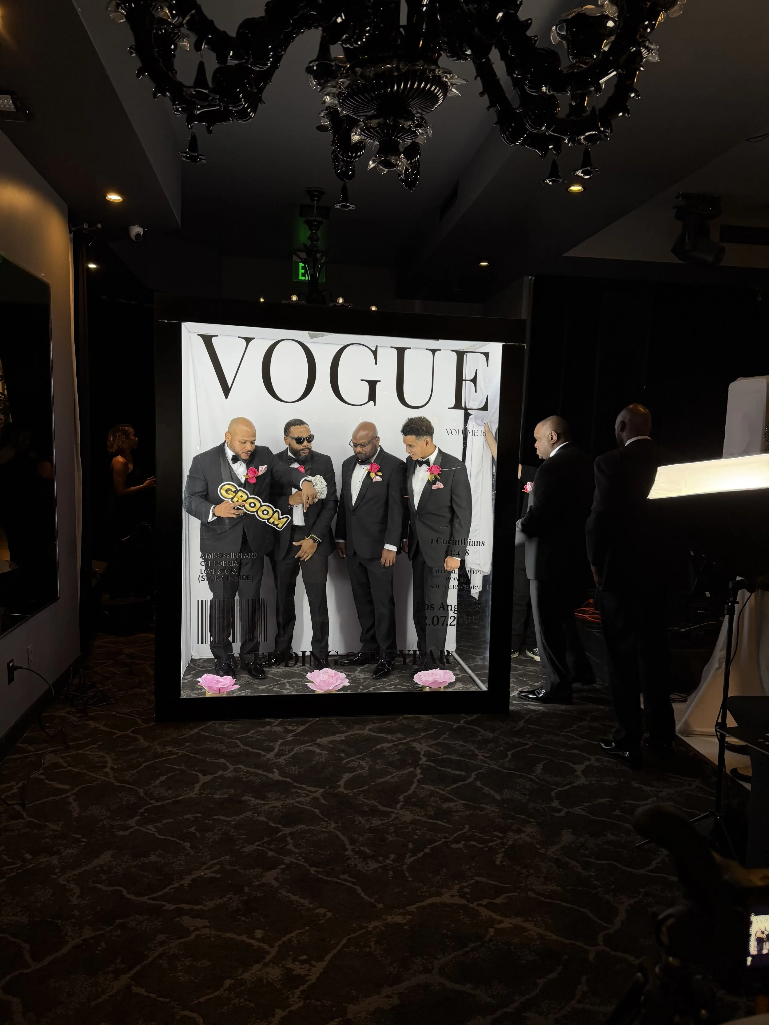 Group of five men in tuxedos with pink boutonnieres posing in front of a Vogue magazine backdrop, some holding props like a 'Groom' sign and bills, at a wedding or special event.