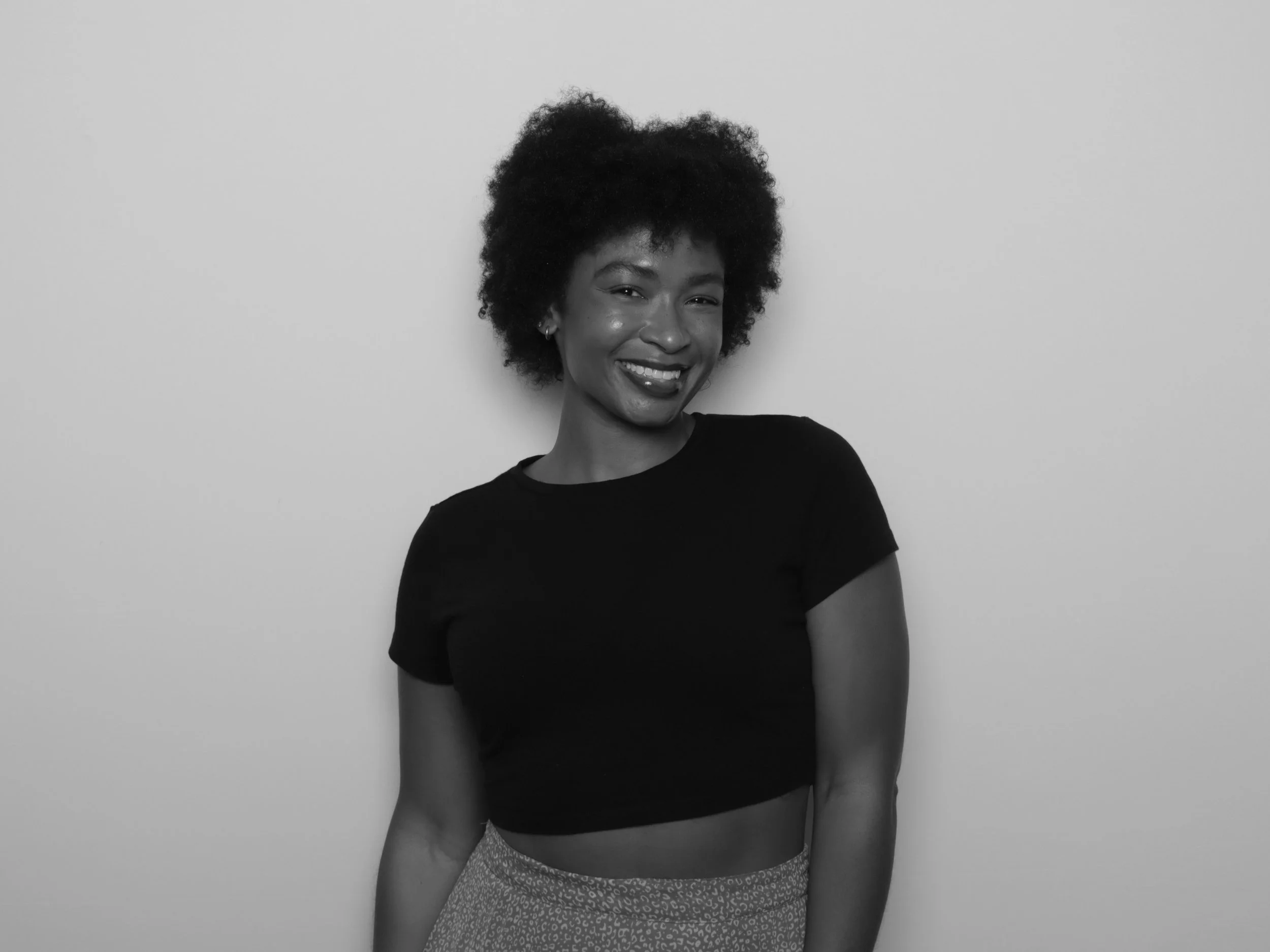 Black and white image of a smiling woman with curly hair, wearing a black t-shirt and patterned skirt, standing against a plain white wall.