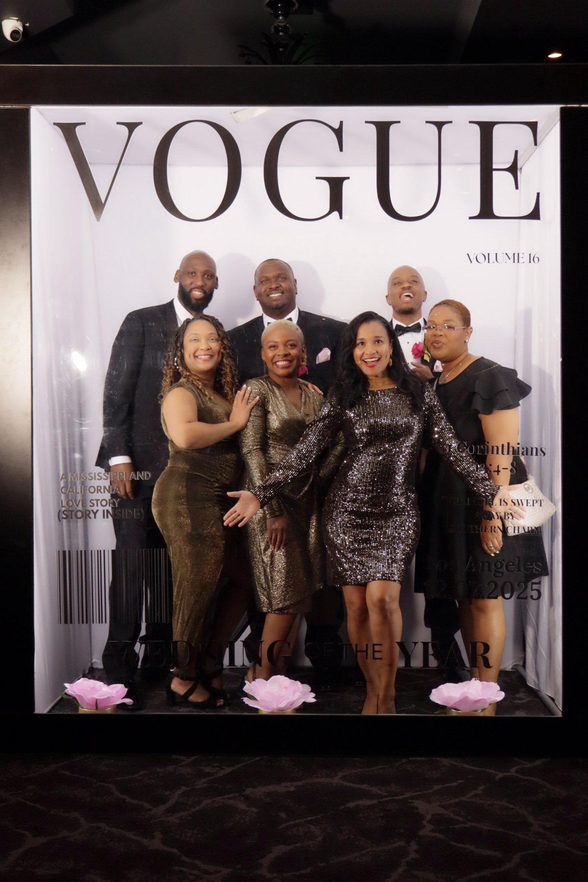 A group of seven people in formal attire posing for a photo at a Vogue event. The group includes four women and three men, standing in front of a large white backdrop with the Vogue logo and event details. They are smiling and appear to be happy.