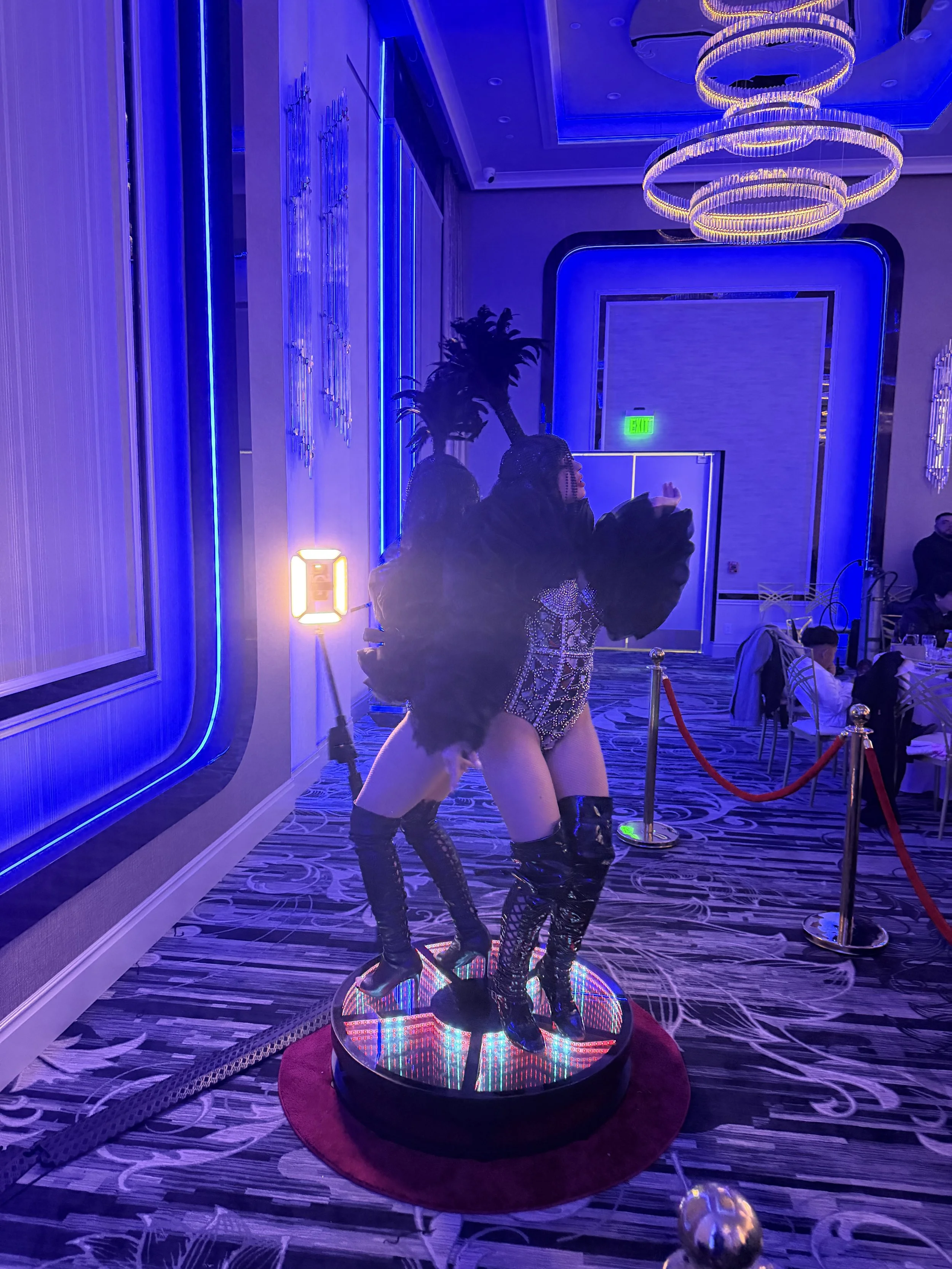 A modern, vibrant dance-floor display featuring a mannequin dressed in elaborate, rhinestone-studded clothing and high boots with an LED light-up platform, situated inside a decorated room with blue and purple lighting, chandeliers, and a carpeted fl