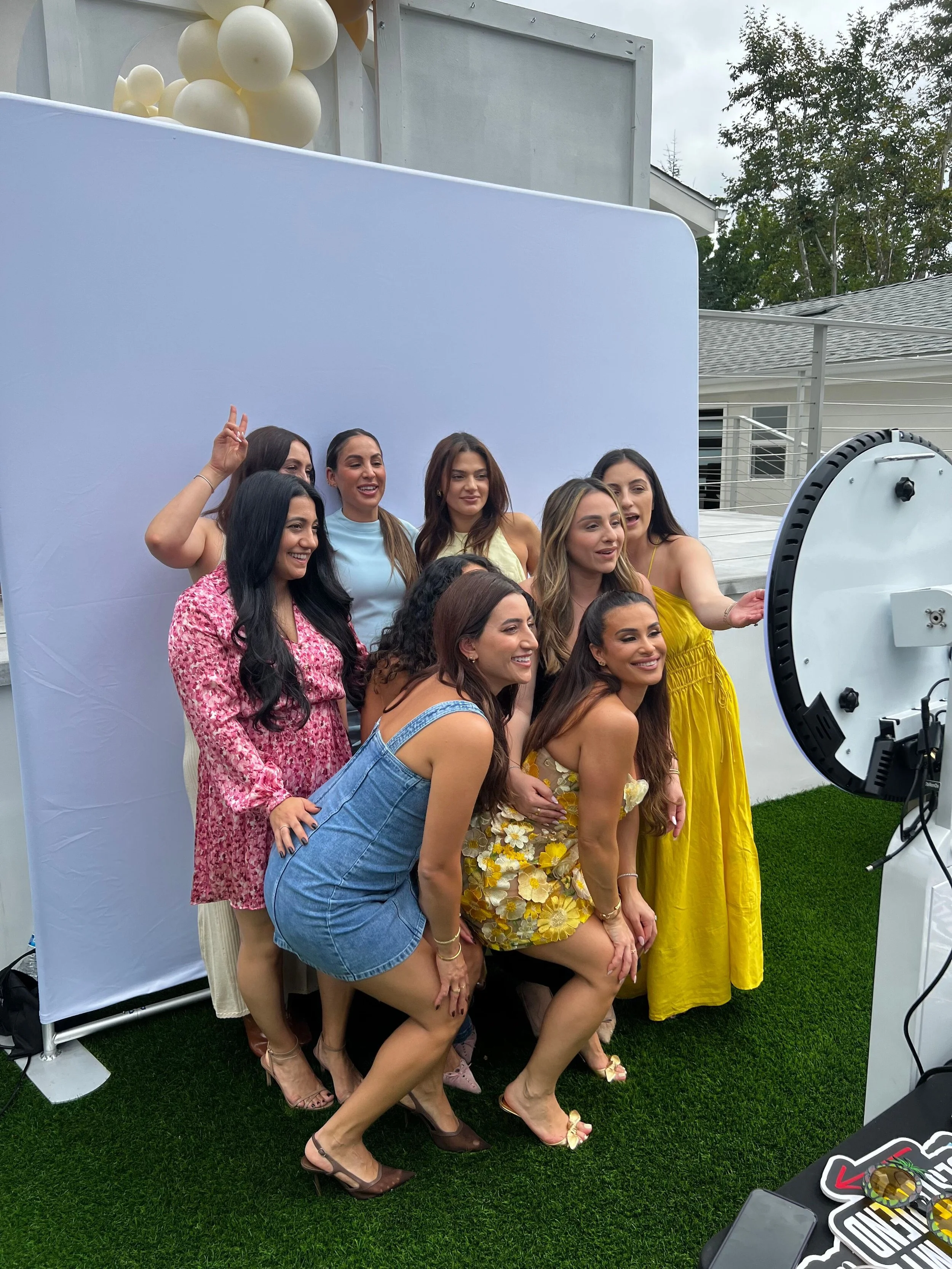 Group of ten women taking a photo together outdoors, some making peace signs, with a blue backdrop and balloons in the background.