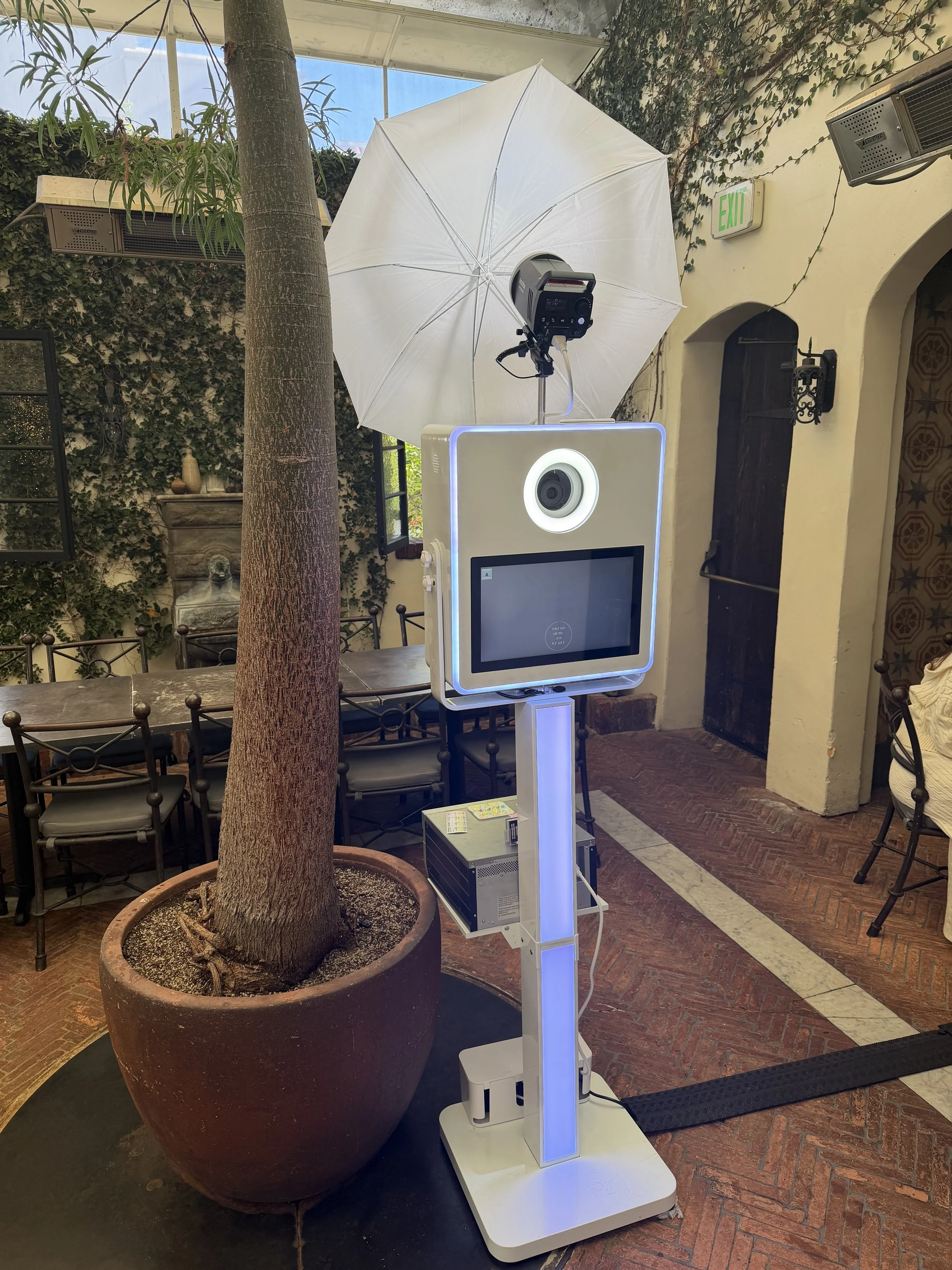 A photo of a photo booth with a large white umbrella, a camera, a large display screen, and a stand. The setting is indoors, with a potted tree and chairs in the background, and an emergency exit sign on the wall.