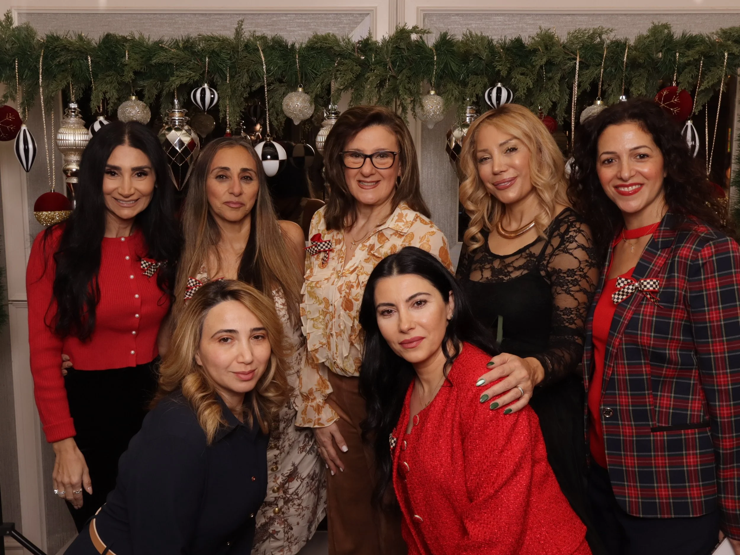 Group of eight women celebrating Christmas indoors with festive decorations and greenery in the background.
