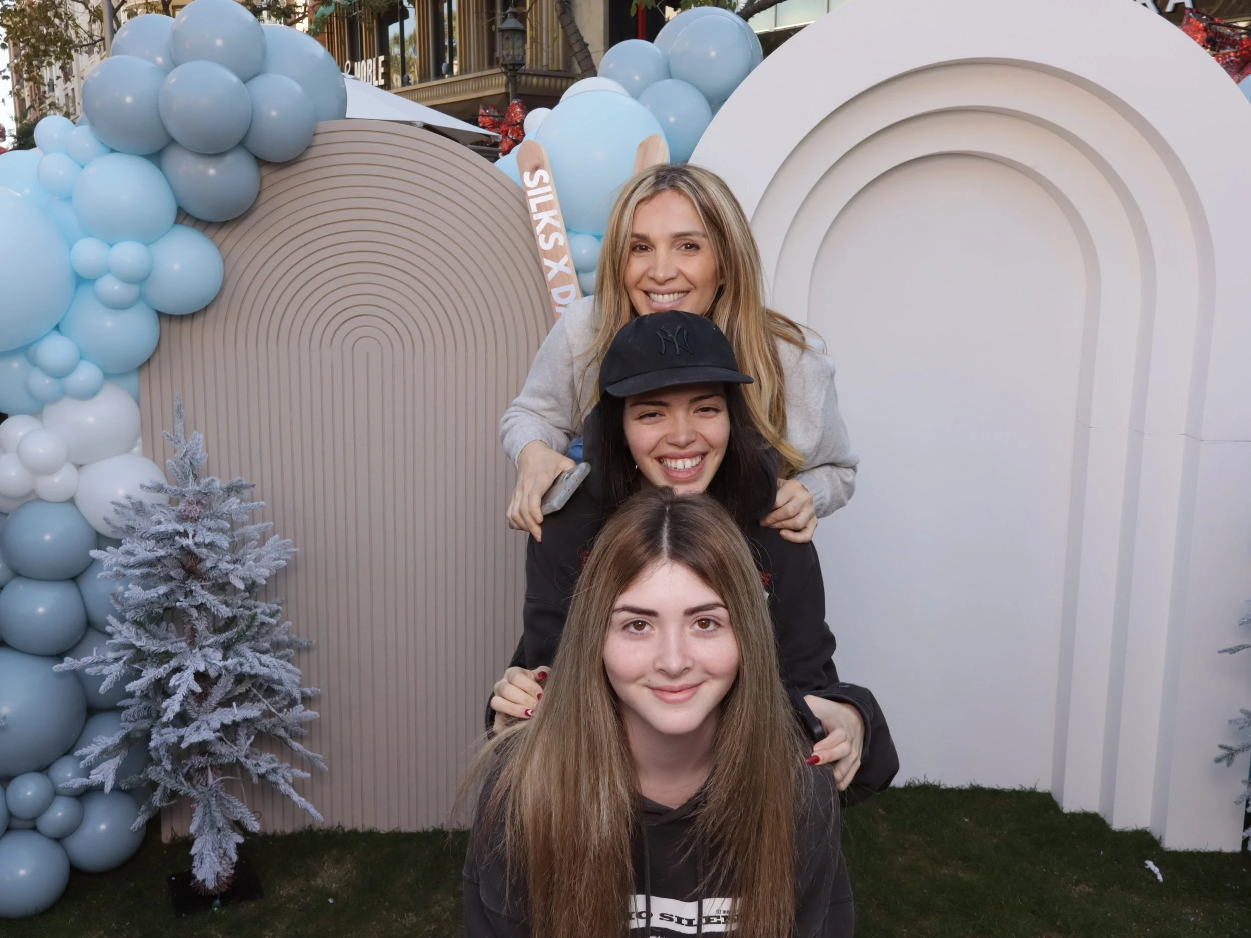 Three women posing in a stacked formation outdoors with festive decorations, including large balloons and Christmas trees, behind them.