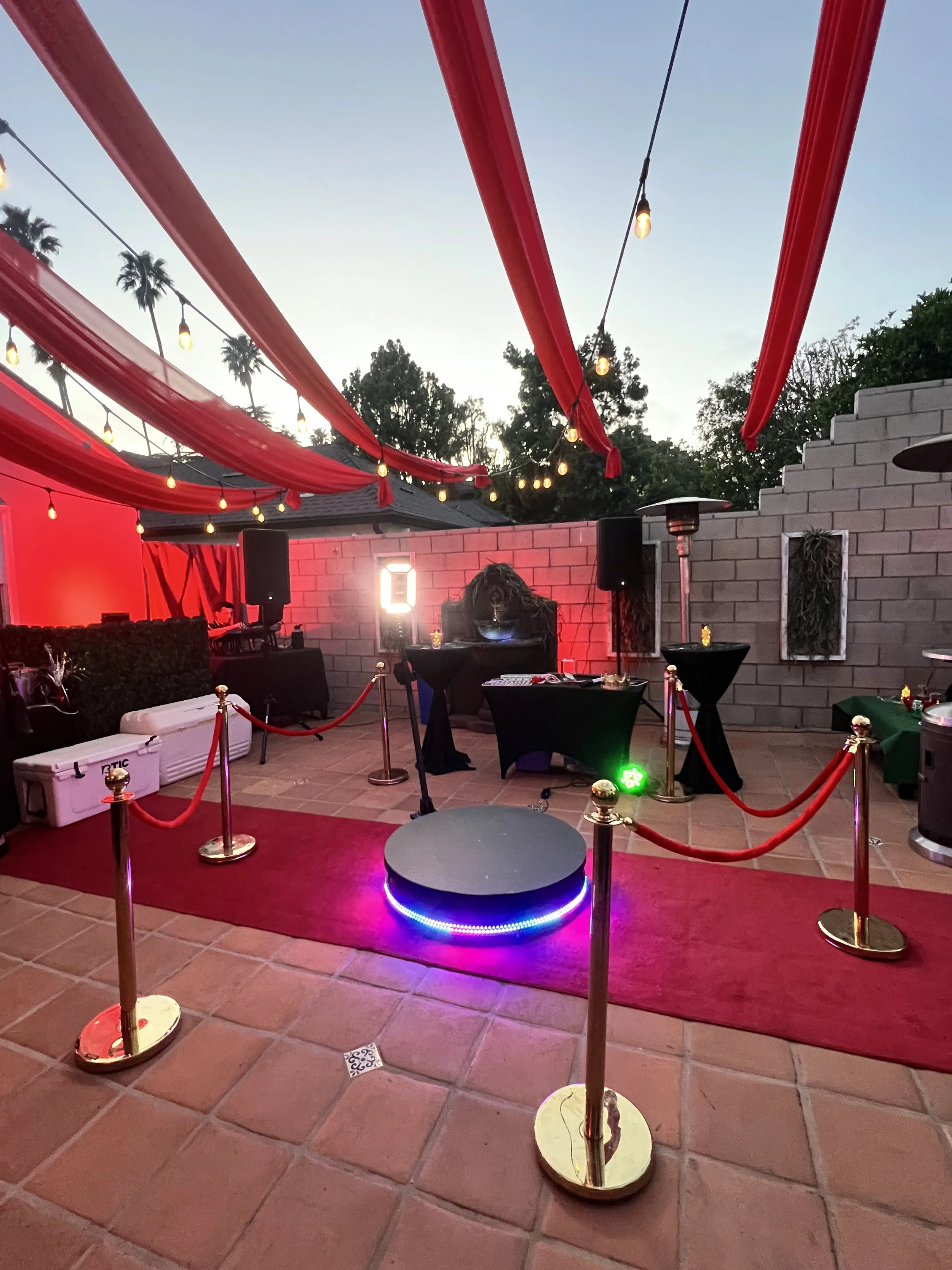Decorated outdoor patio area with red drapes, string lights, a red carpet, and a DJ setup with turntable and speakers, suggesting a celebration or event at sunset.
