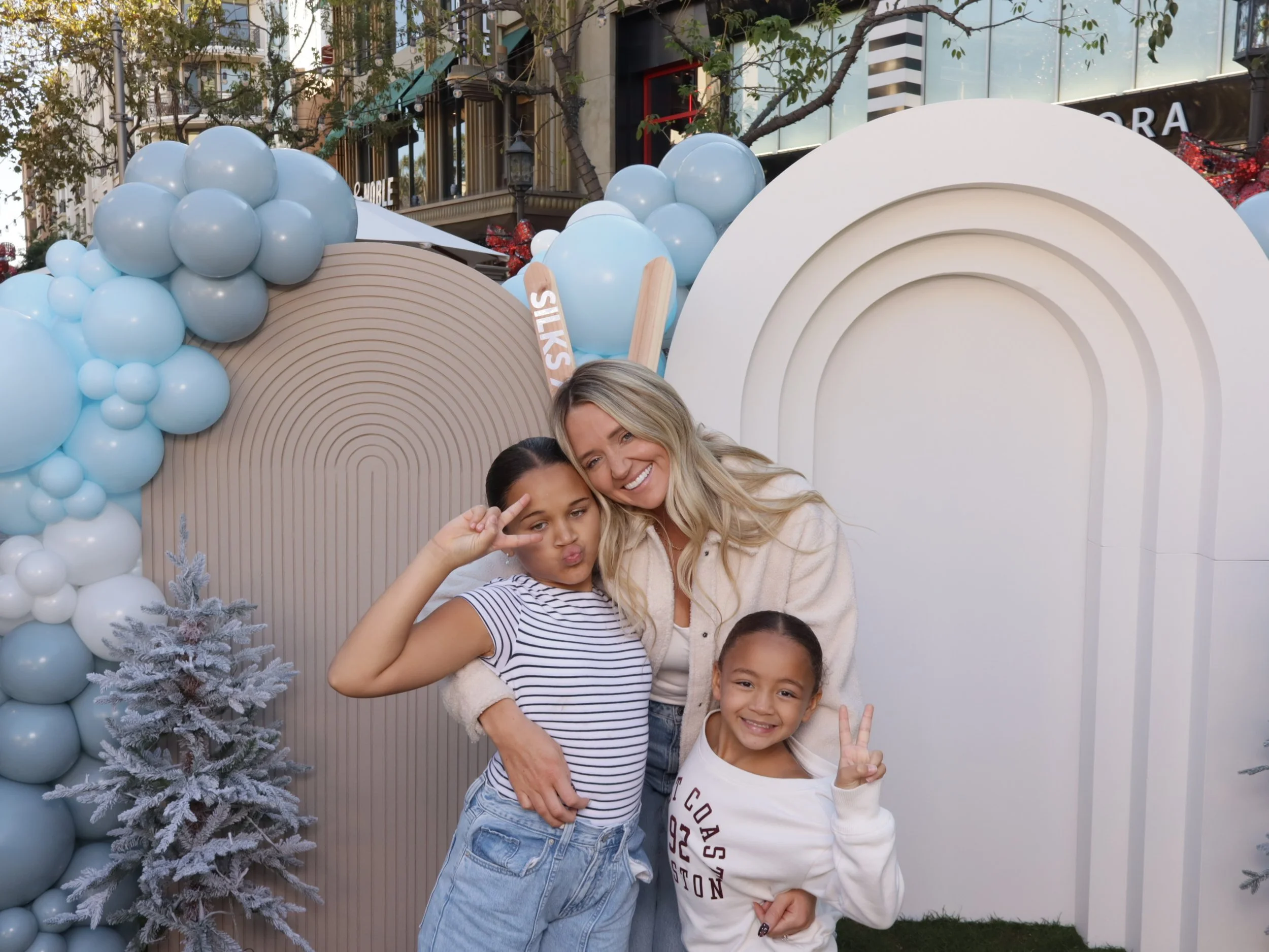 Two young girls and a woman posing with peace signs in front of holiday-themed decorations with balloons and artificial trees.