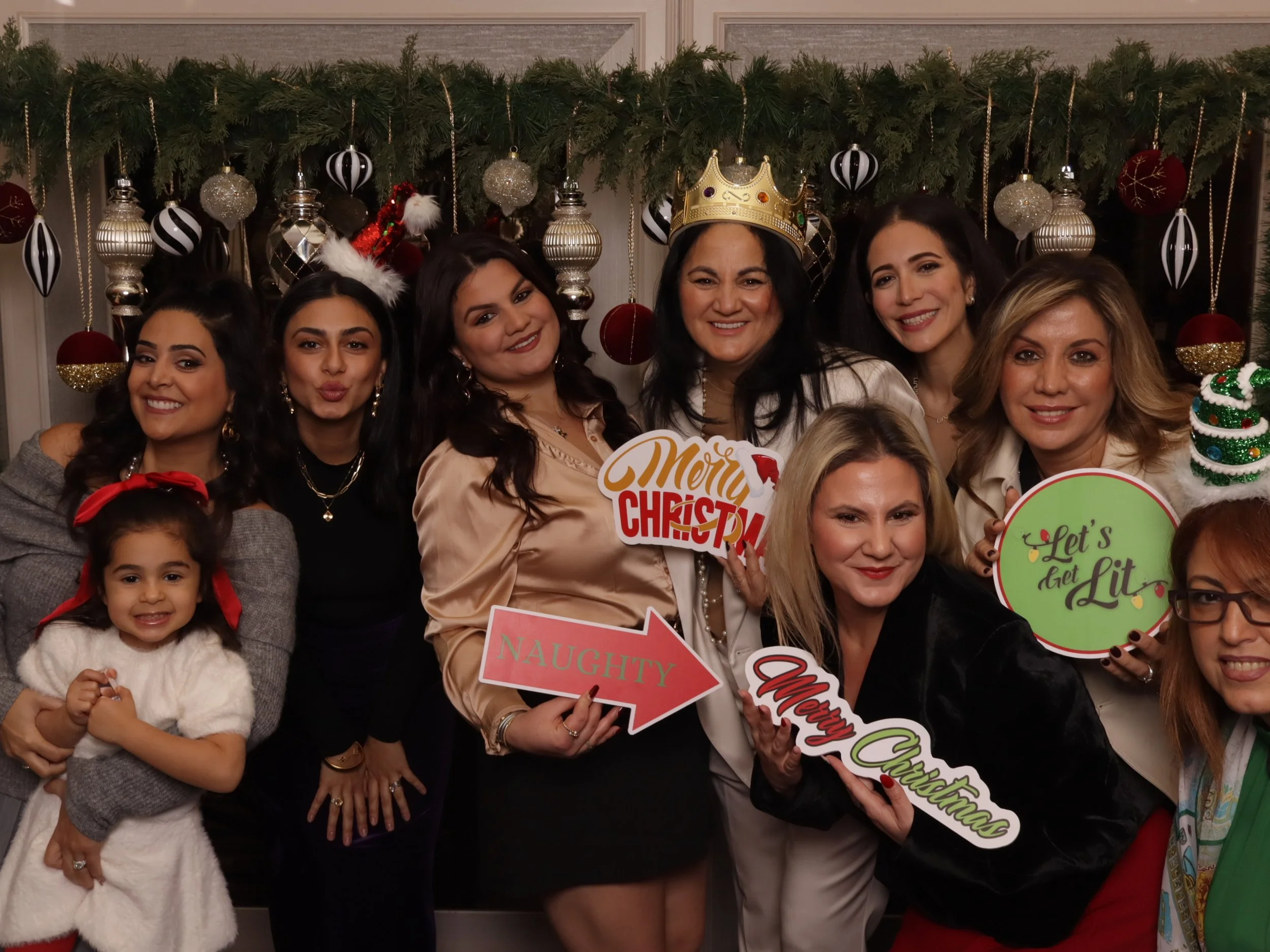 A group of women and a young girl celebrating Christmas indoors. They are holding festive signs, including one that says 'Merry Christmas,' and are wearing holiday accessories. The background features Christmas decorations with ornaments and greenery