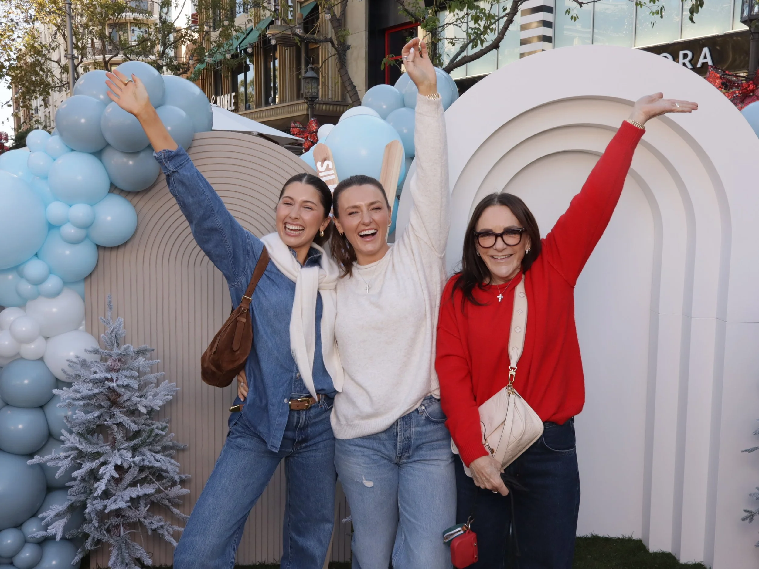 Three women smiling and celebrating outdoors at a festive event with holiday decorations, including blue and white balloons and frosted trees.