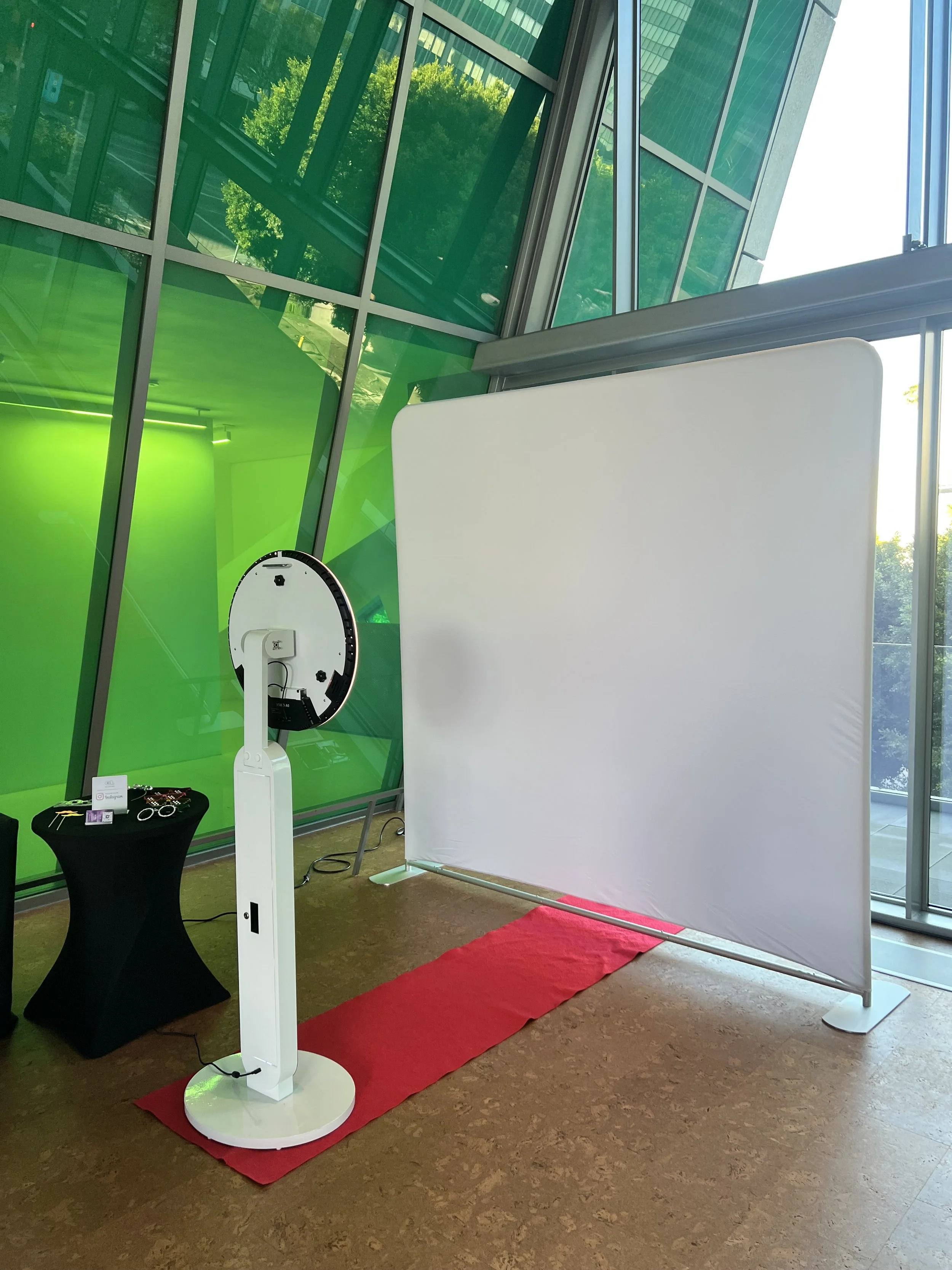 Photo of a photo booth setup inside a modern building with large glass windows and green walls. The booth includes a white background, a stand with a circular camera or scanner, and a small black table with items on it. There is a red carpet in front