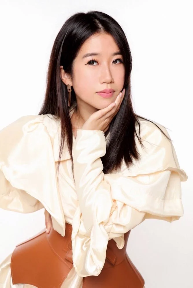 A woman with long black hair, wearing a cream-colored top and tan skirt, posing with her hand on her chin against a plain white background.