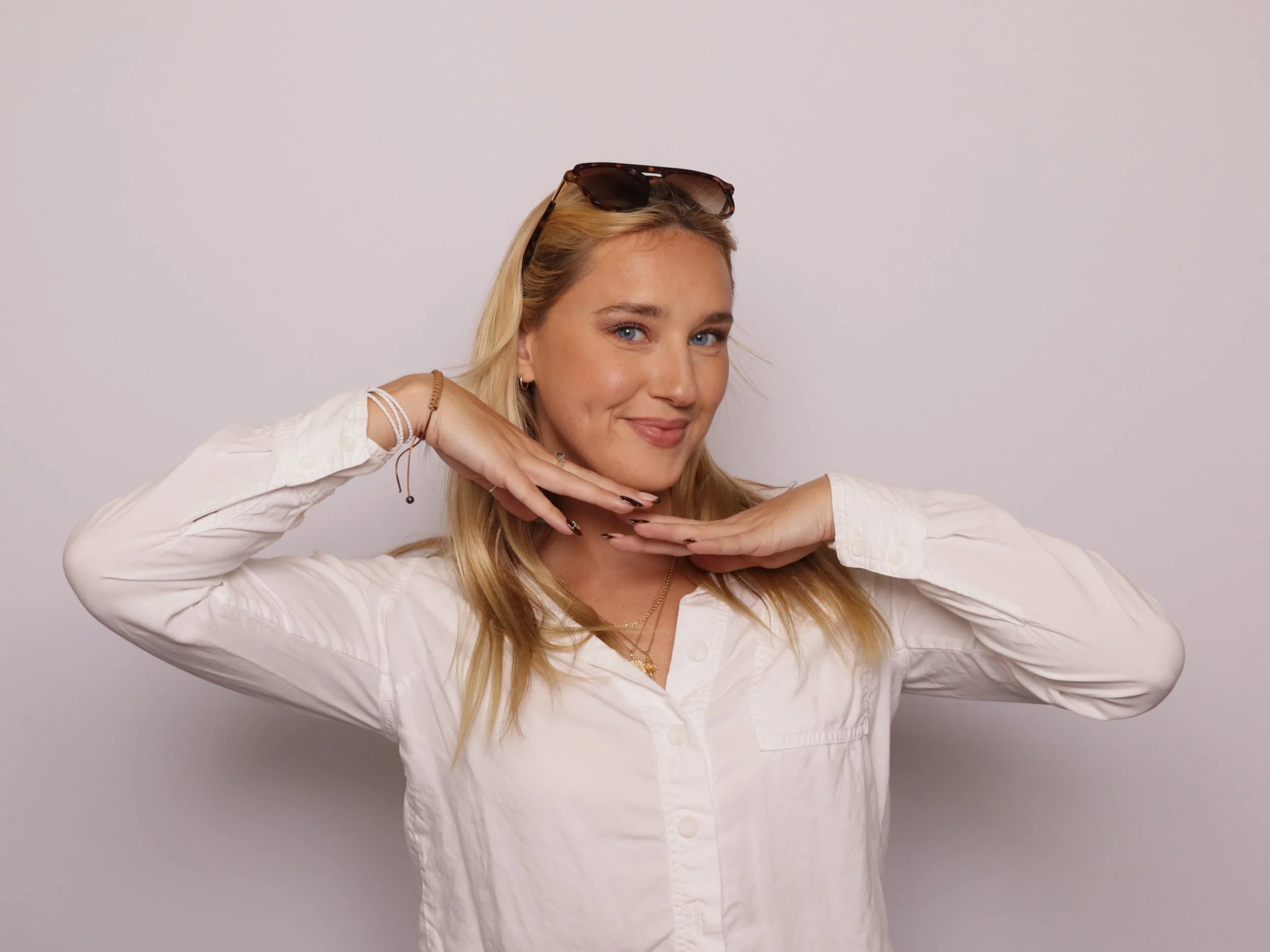 A young woman with blonde hair and blue eyes, wearing a white button-up shirt and sunglasses on her head, poses with her hands under her chin against a gray background.