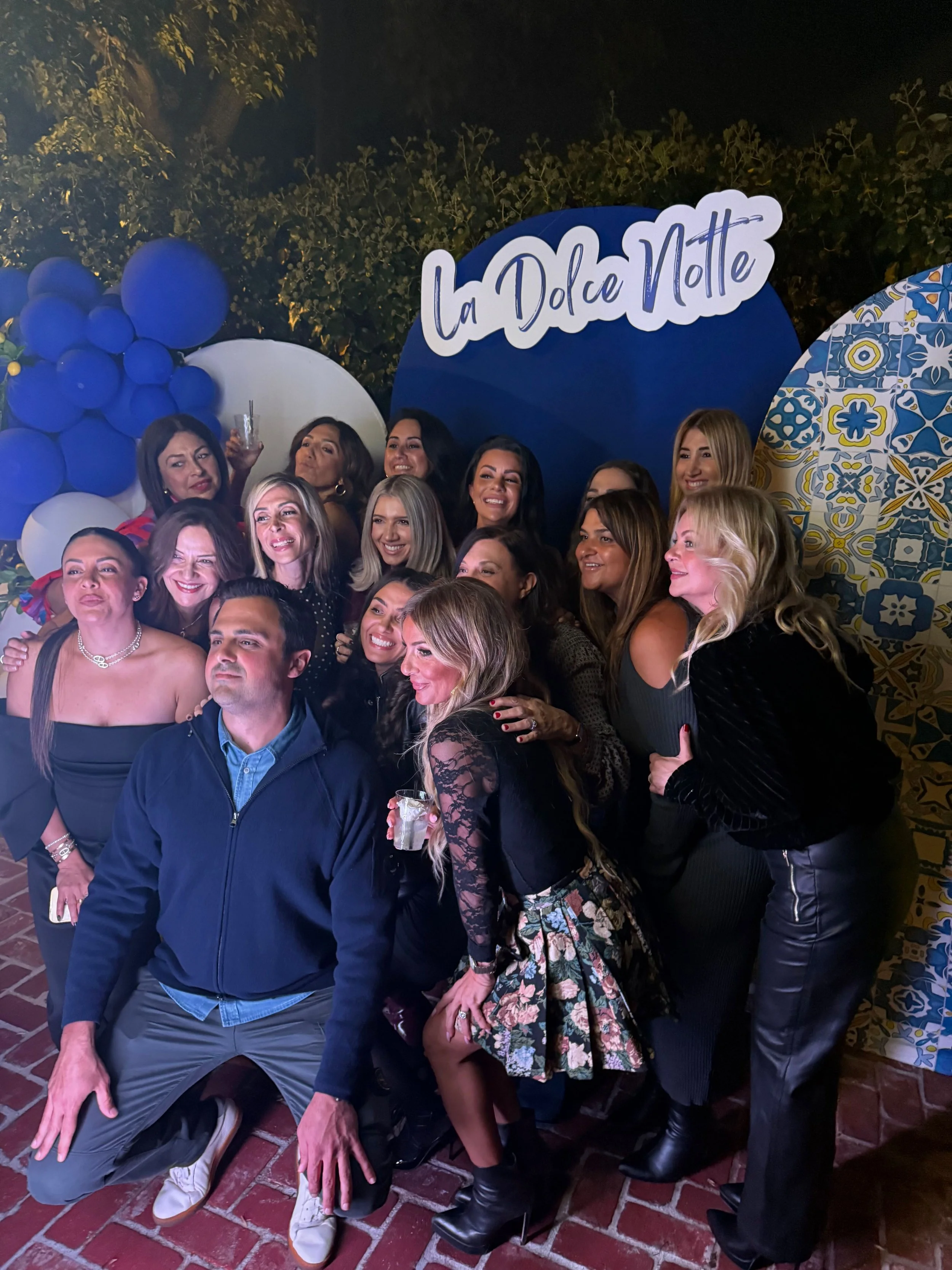 Group of women and one man posing together at an event with a 'La Dolce Vita' sign, balloons, and decorative backgrounds.