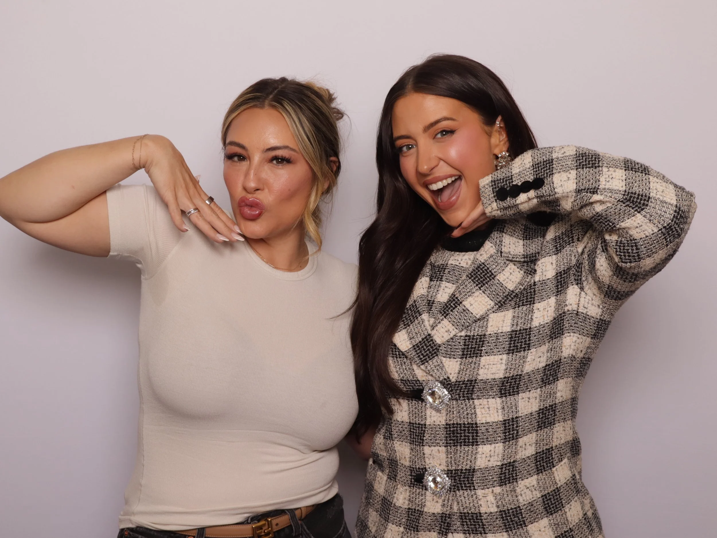 Two women posing playfully in front of a plain white background, with one making a kissy face and the other smiling widely.