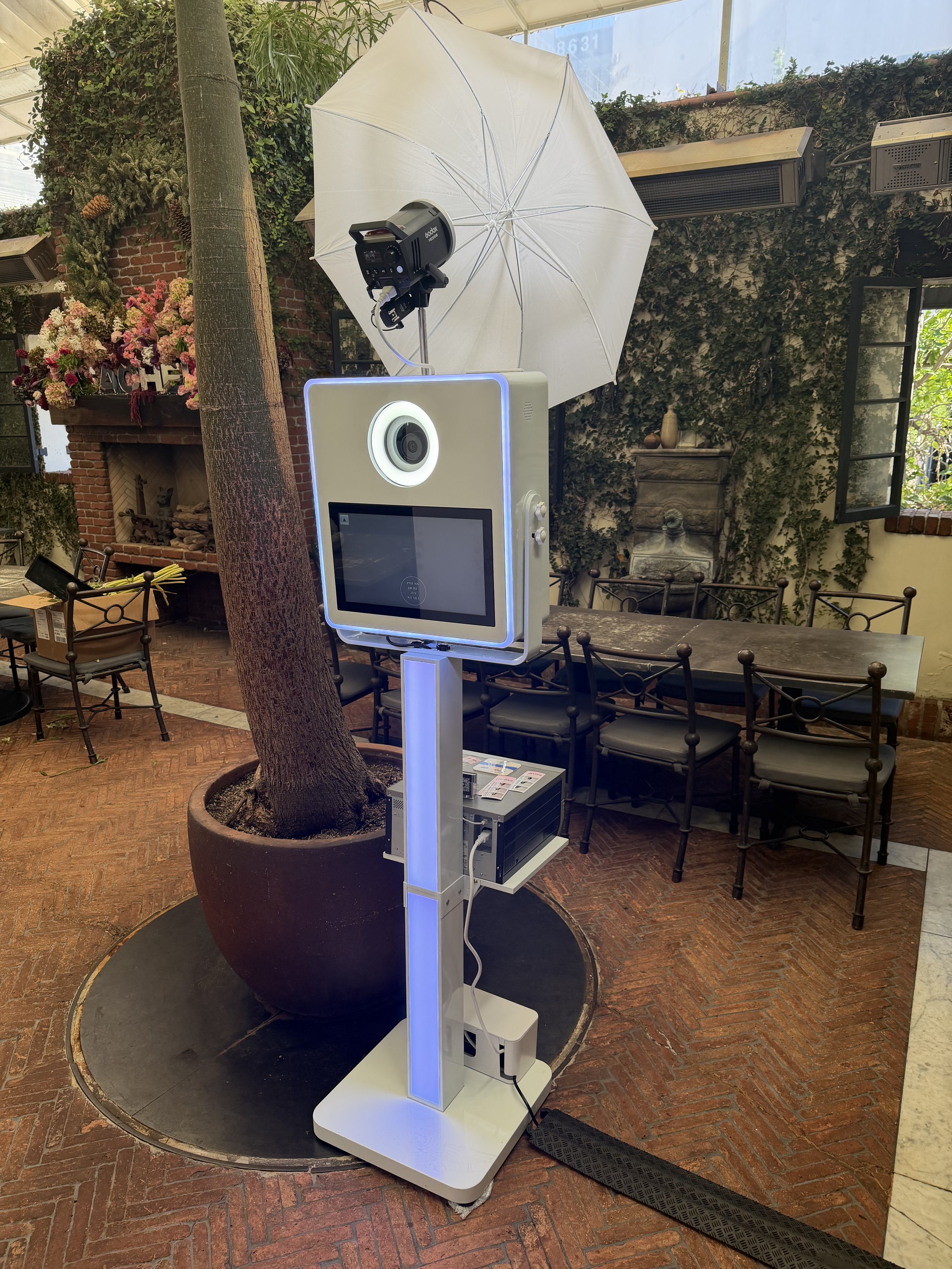 A photo of a photobooth setup with a large white umbrella, camera on top, a touchscreen interface, and a light strip. The setup is indoors, near a potted tree and a brick fireplace with floral decorations, in a rustic-style dining area.