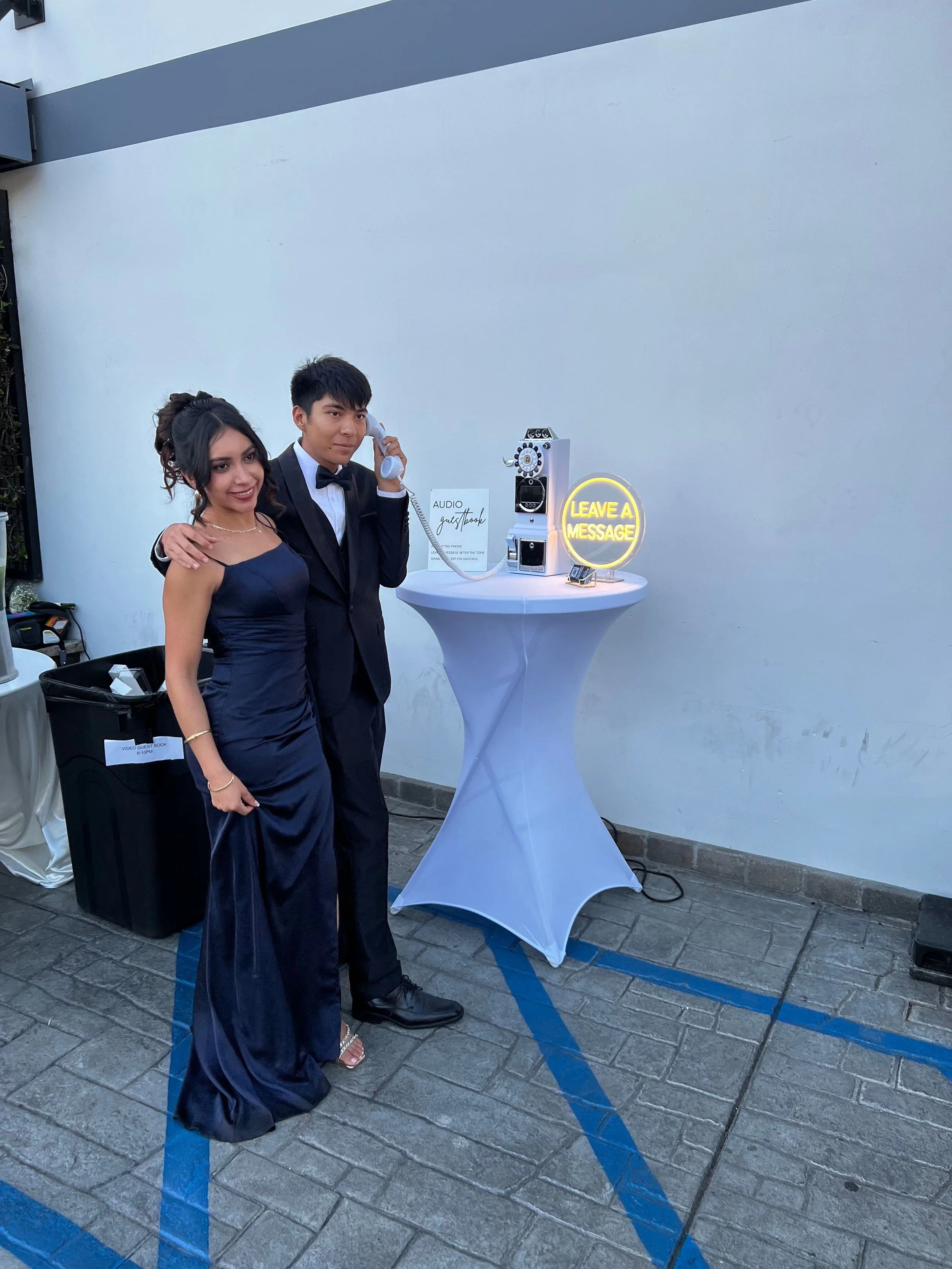 A young woman in a dark blue evening gown and a young man in a tuxedo with a bow tie stand together, smiling, at a photo booth station with a vintage payphone, a neon sign saying 'Leave a Message,' and a sign that reads 'Audio Guest Book.'