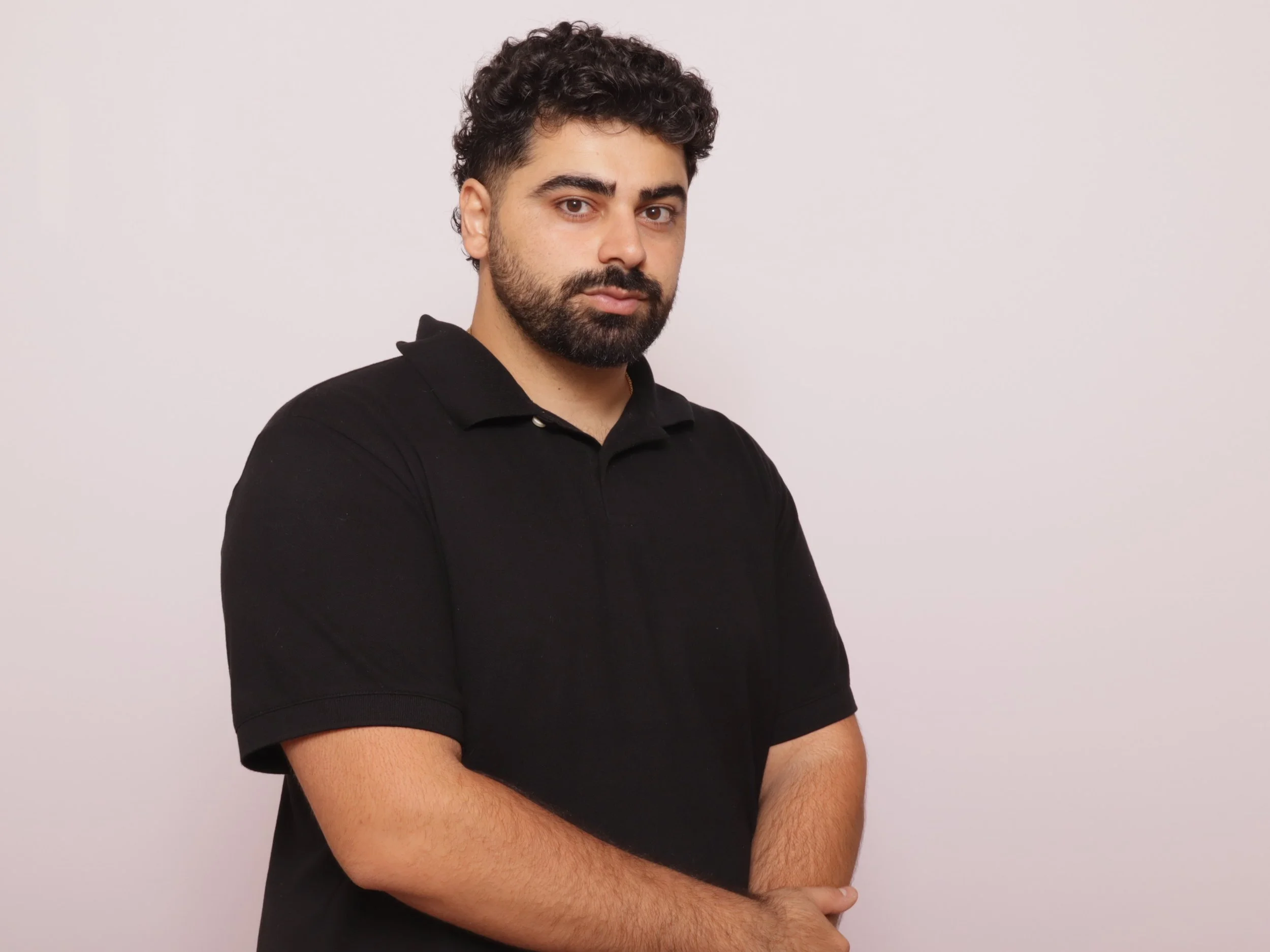 A man with dark curly hair, beard, and mustache wearing a black polo shirt standing against a plain white background.