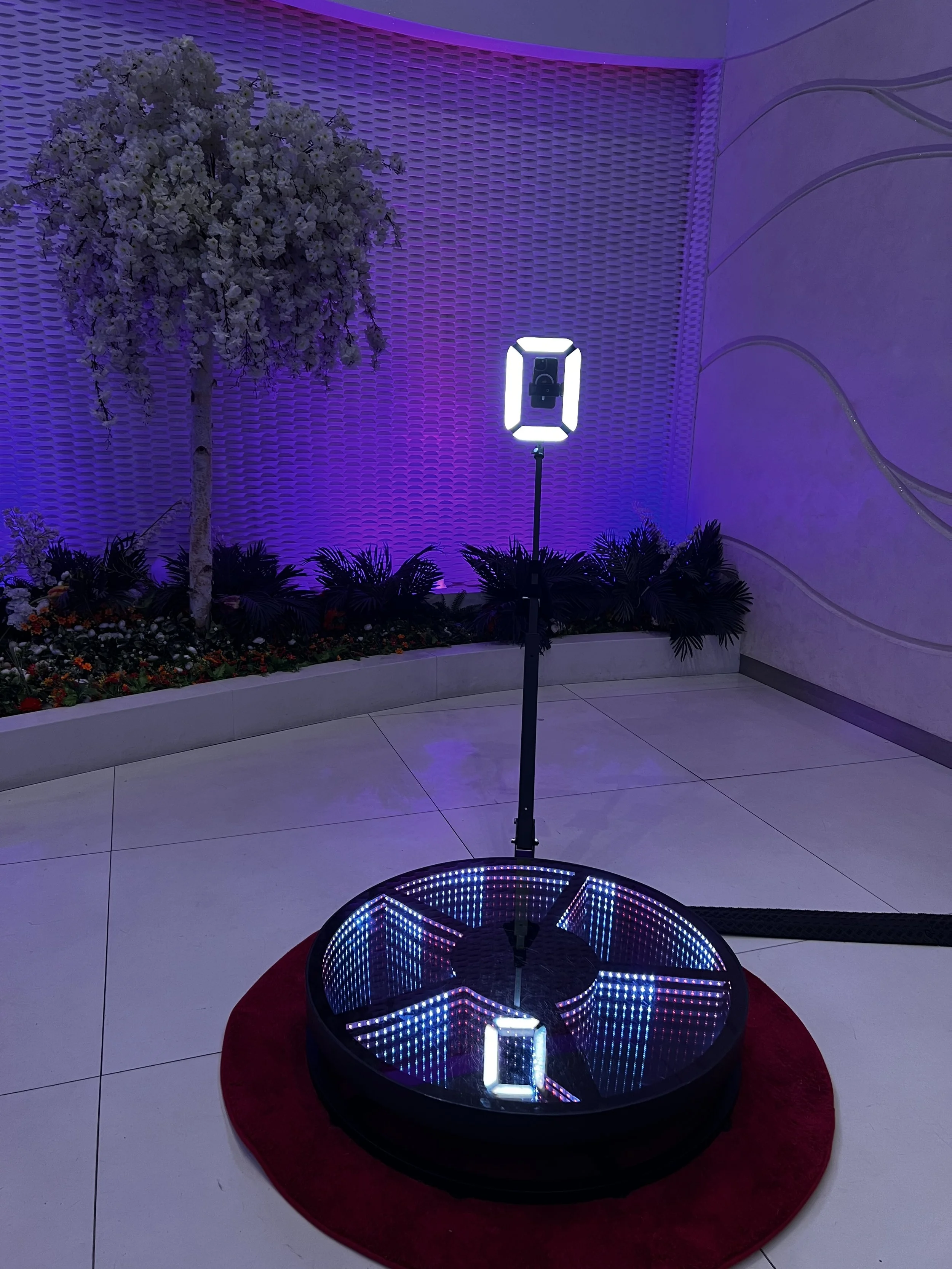 A dance floor with LED lights and a large mirror at the center, set in an indoor venue with purple lighting, a decorative flower arrangement, and a textured wall in the background.