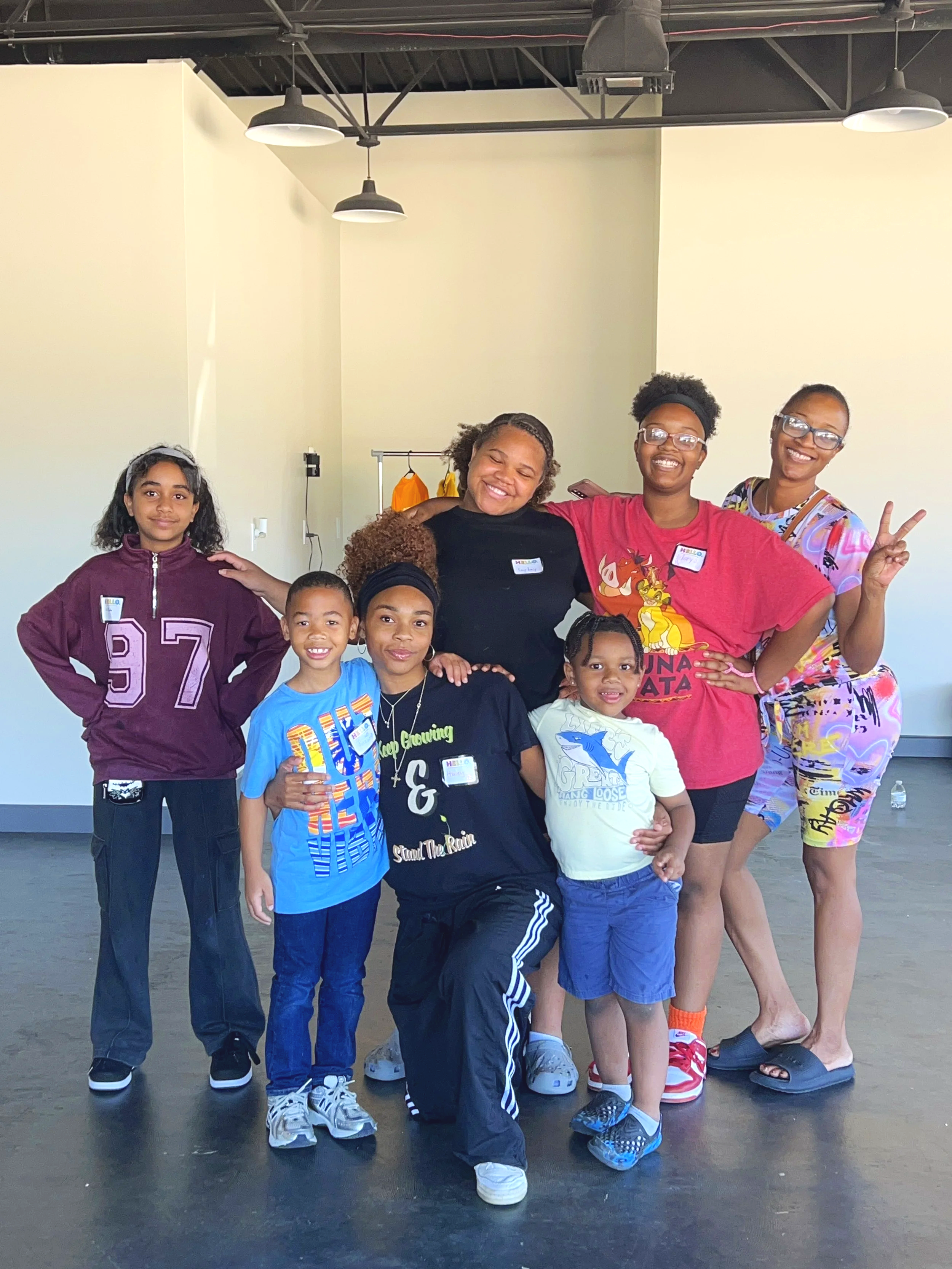 Group of six women and children, smiling and posing together indoors. The group includes three young children and three women, some wearing glasses. They are standing against a plain wall with the ceiling and lighting visible.
