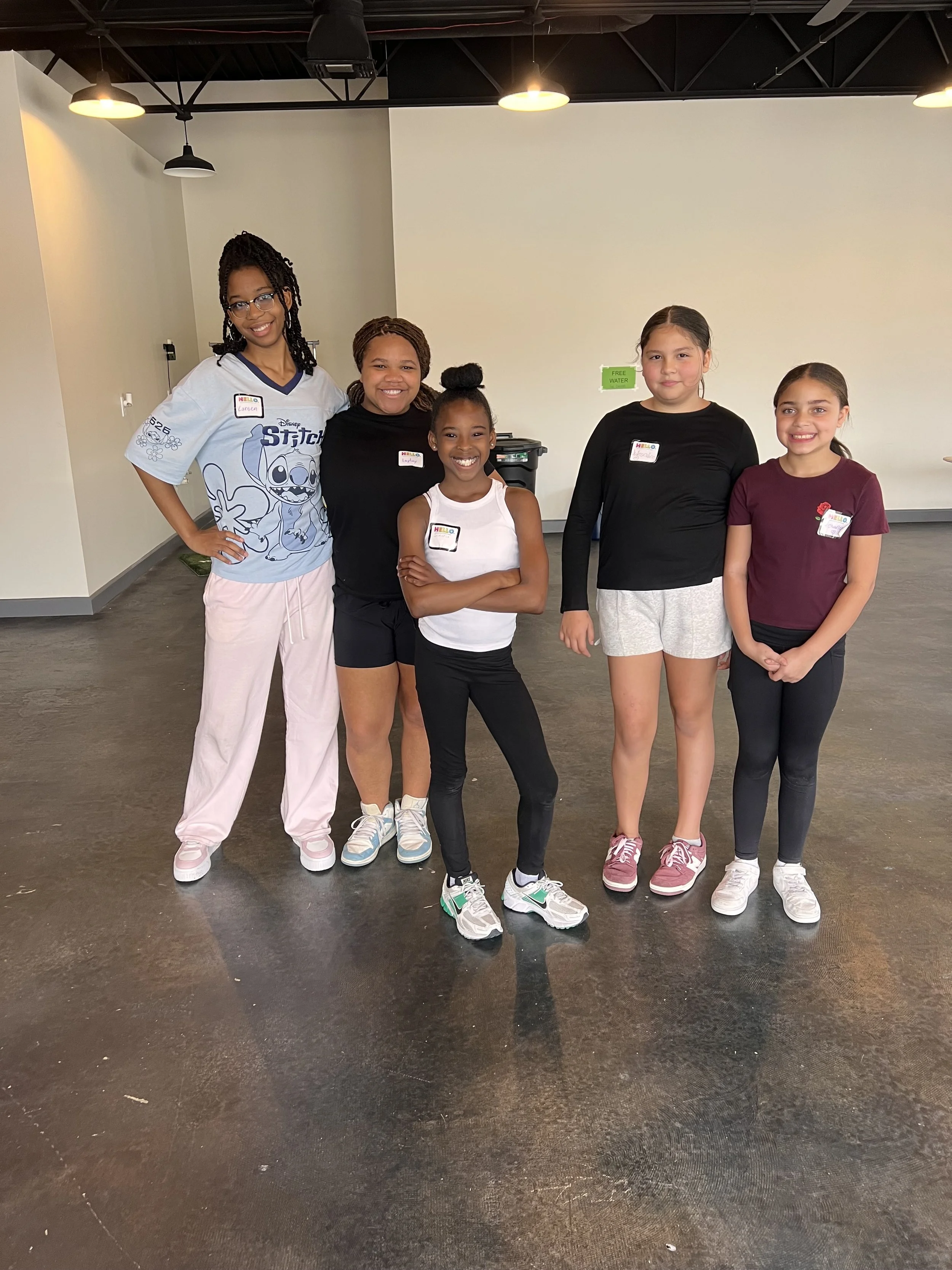 Group of five young girls standing indoors, smiling at the camera.