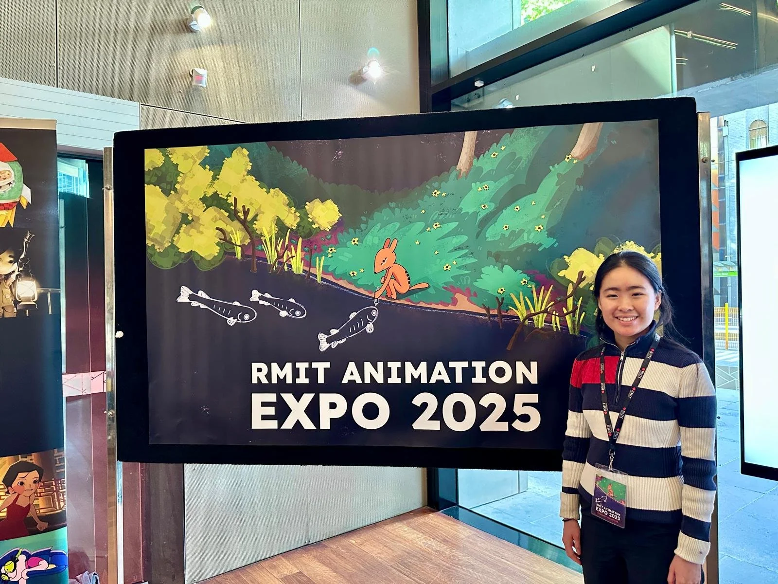 A smiling woman standing next to a sign for the RMIT Animation Expo 2025. The sign features an animated scene with a rabbit, a stream with fish, trees, and greenery.