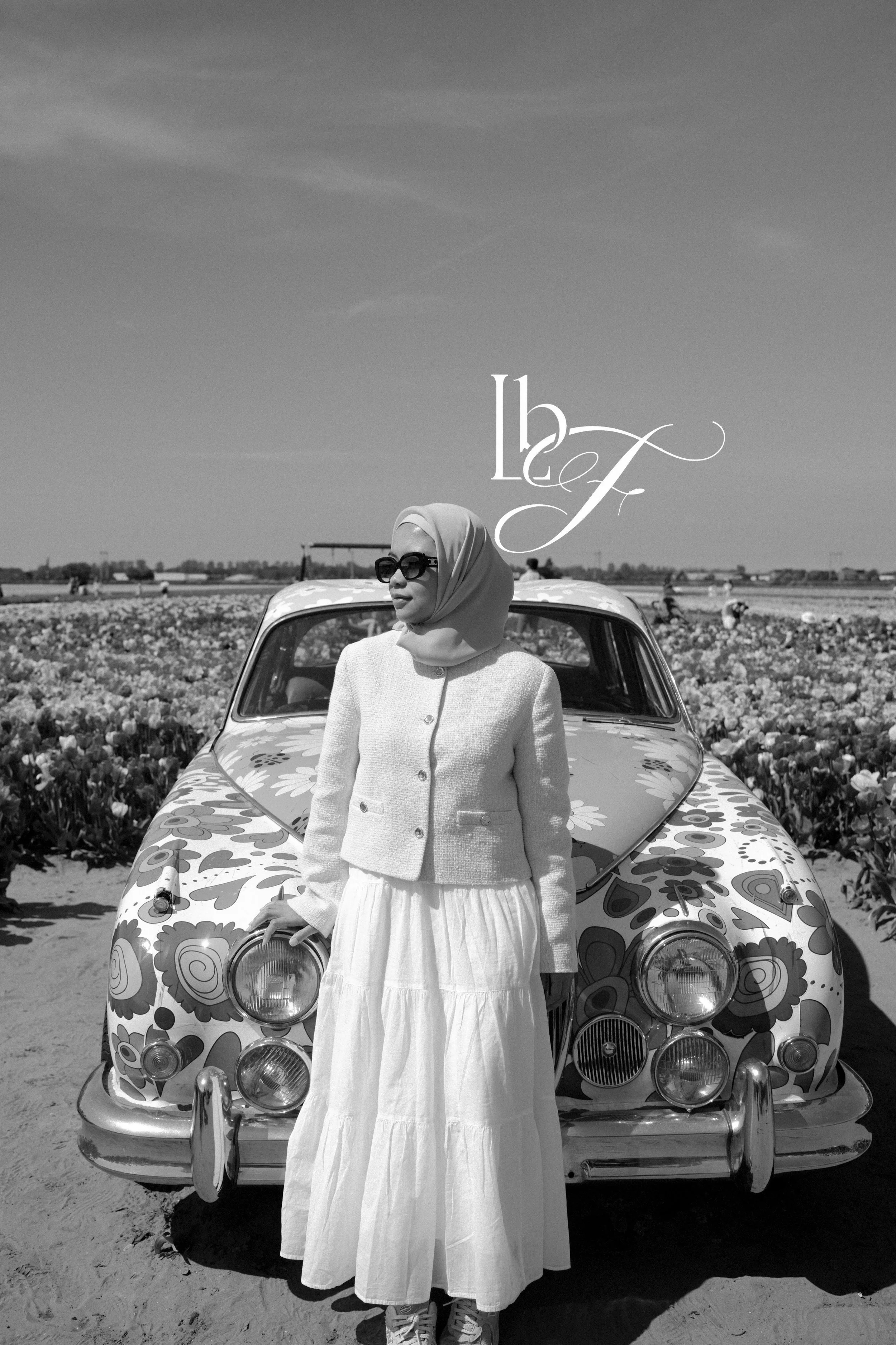 A woman wearing sunglasses, a hijab, a light-colored jacket, and a long skirt standing in front of a vintage floral-patterned car in a field of flowers on a sunny day.