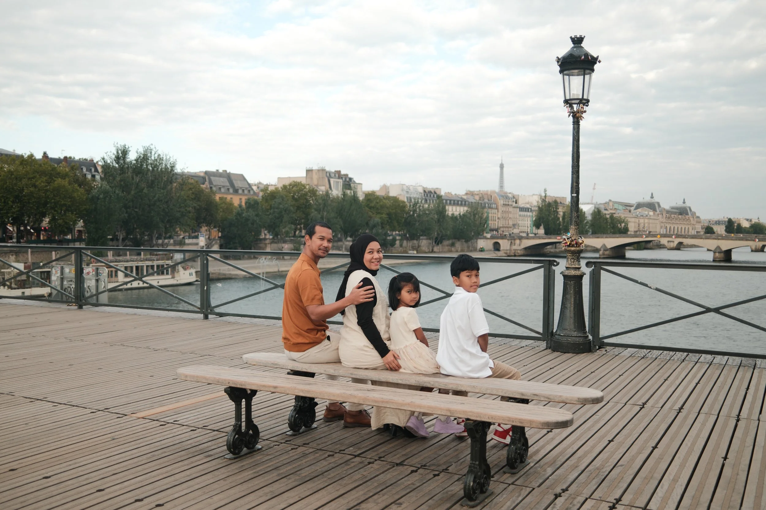 A family of four sitting on a bench by the river in Paris, with the Eiffel Tower visible in the background.