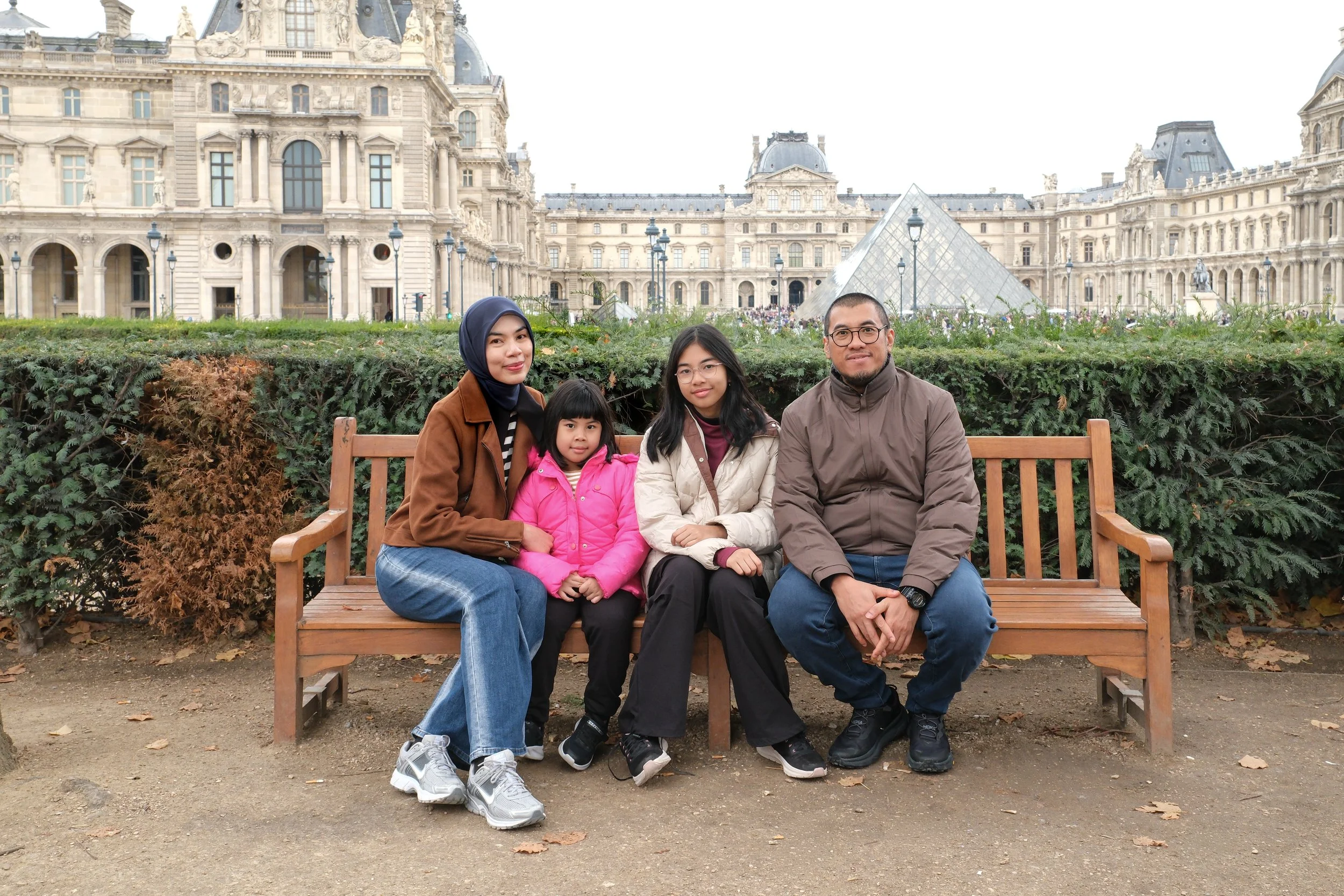 Family of four sitting on a park bench in front of the Louvre Museum in Paris, France. The family includes a woman in a hijab, a girl in a pink jacket, a woman in a beige jacket, and a man in a brown jacket. The Louvre Pyramid is visible in the backg